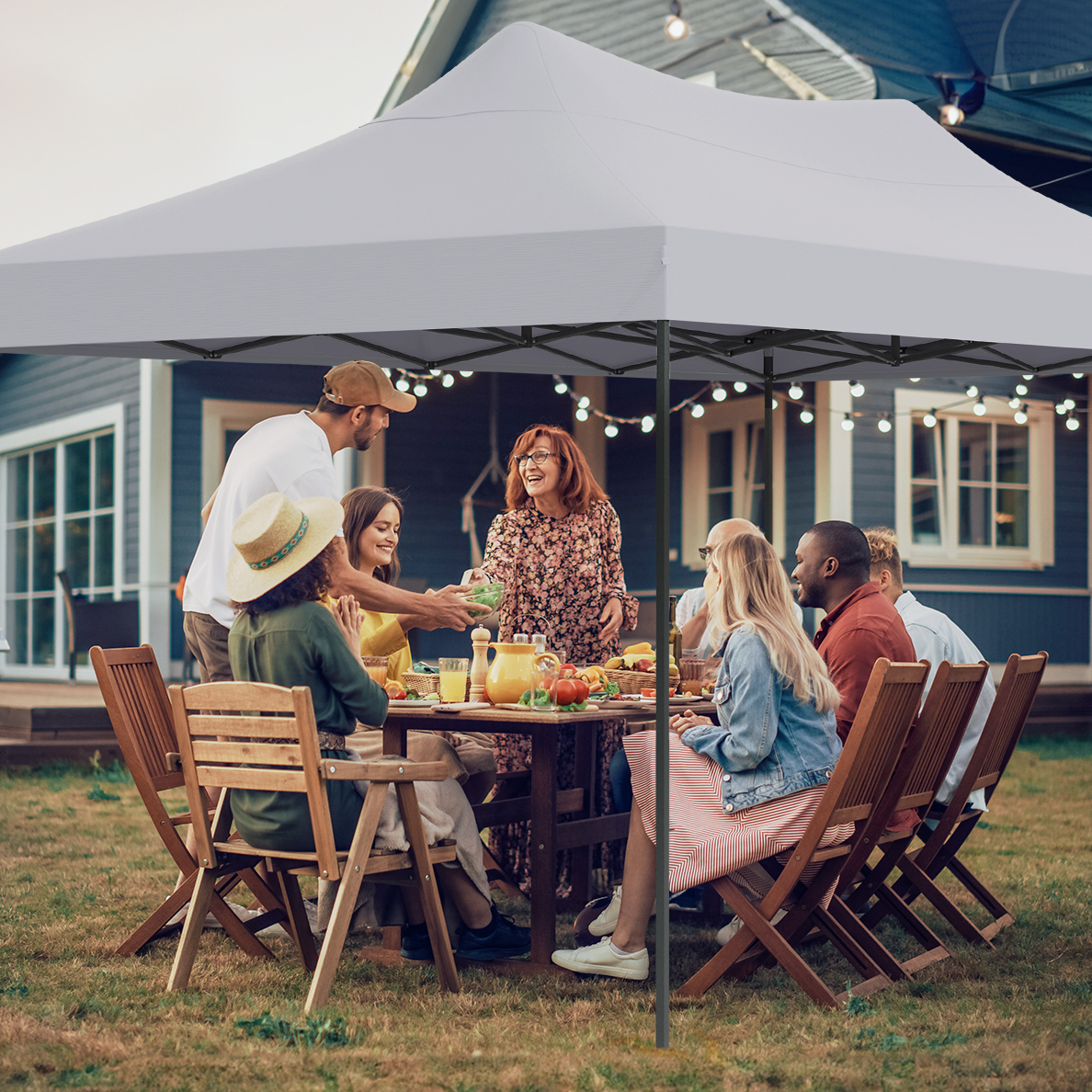 A group of people are seated around a table under a white tent. A man is serving food. A house is in the background.