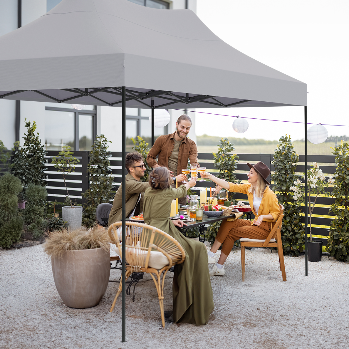 Group of friends toasting with drinks under a canopy in a garden. Table set with food. White gravel. Green plants and a house in the background.