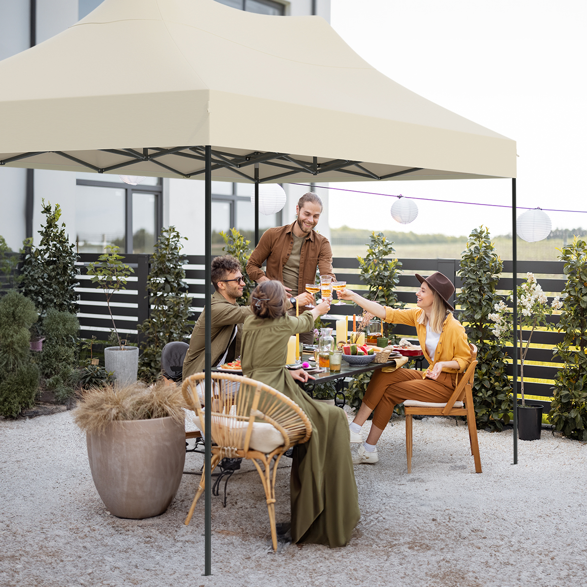 Group of people toasting drinks under a beige canopy in a backyard. They are seated around a table. Decorative lanterns are present.