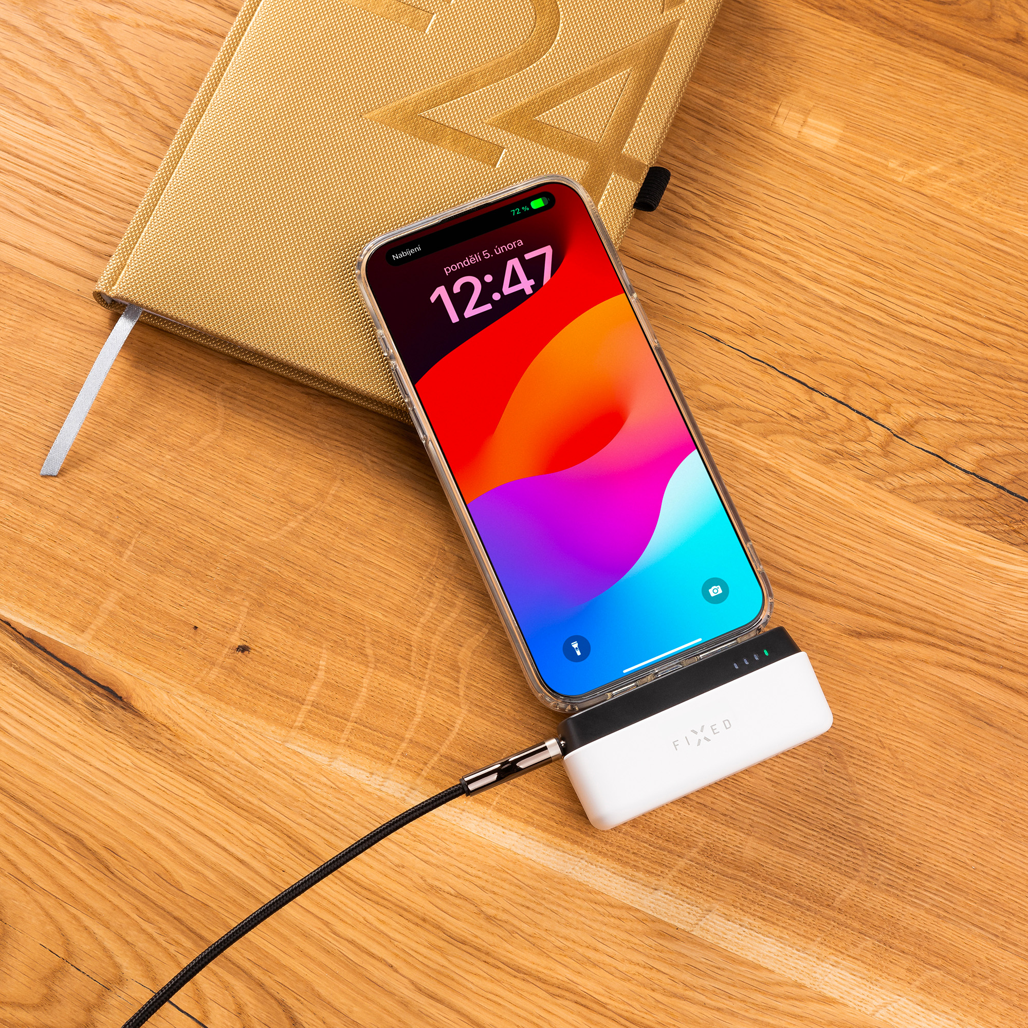 Ein Handy, das auf einem Holztisch geladen wird. Ein goldenes Notizbuch ist sichtbar. Eine Powerbank ist verbunden.