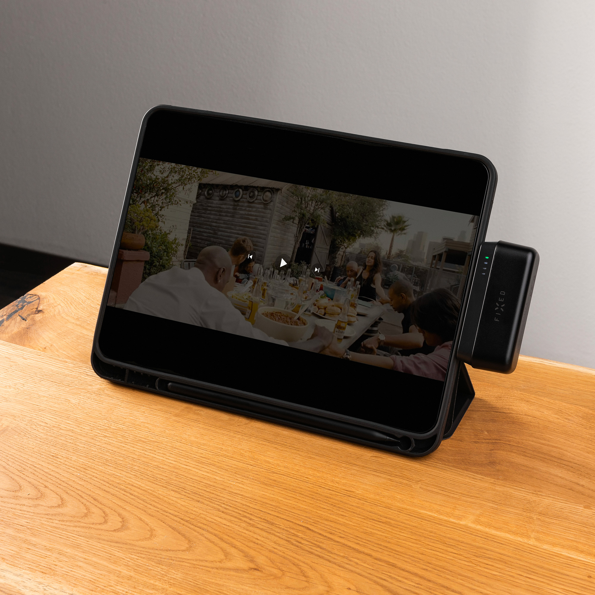 Ein Tablet zeigt ein Video von Personen an einem Tisch. Ein Ladegerät ist am Tablet befestigt, das auf einem Holztisch liegt.