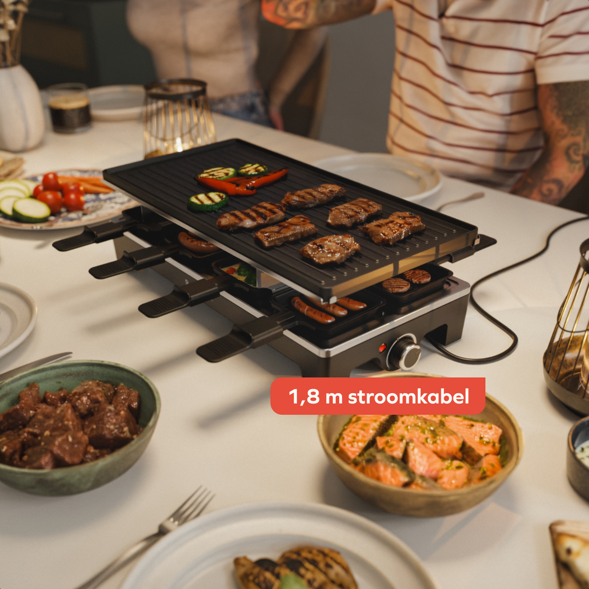 Ein Raclette-Grill mit Fleisch und Gemüse, umgeben von Schüsseln mit Essen, auf einem weißen Tisch. Im Hintergrund sind Personen zu sehen.