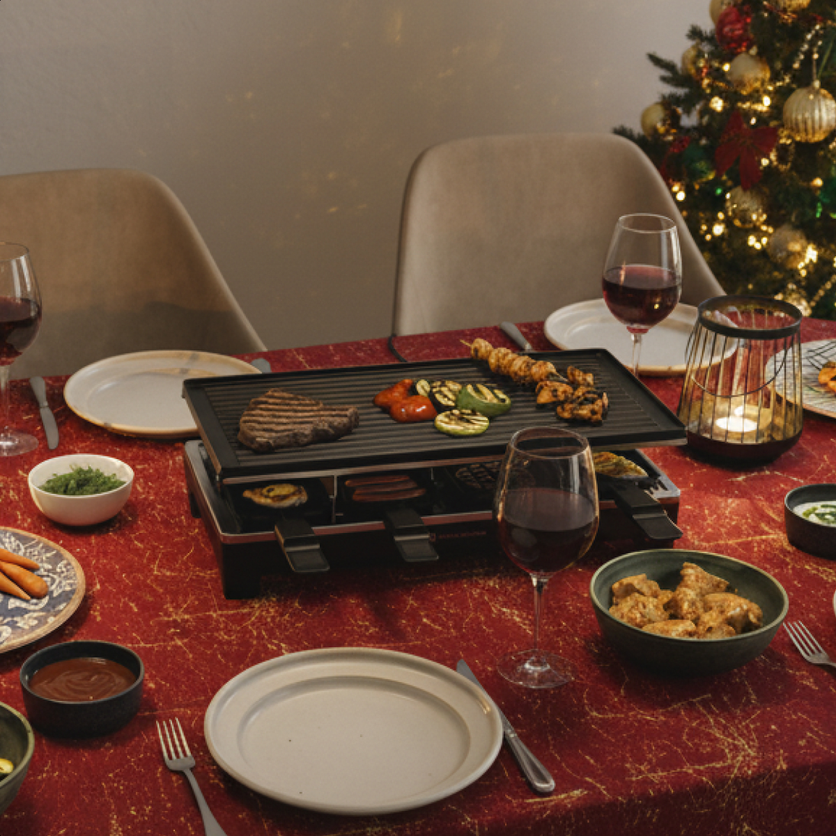 Festlich gedeckter Tisch mit Raclette-Grill, gegrilltem Essen, Weingläsern und einem Weihnachtsbaum im Hintergrund.