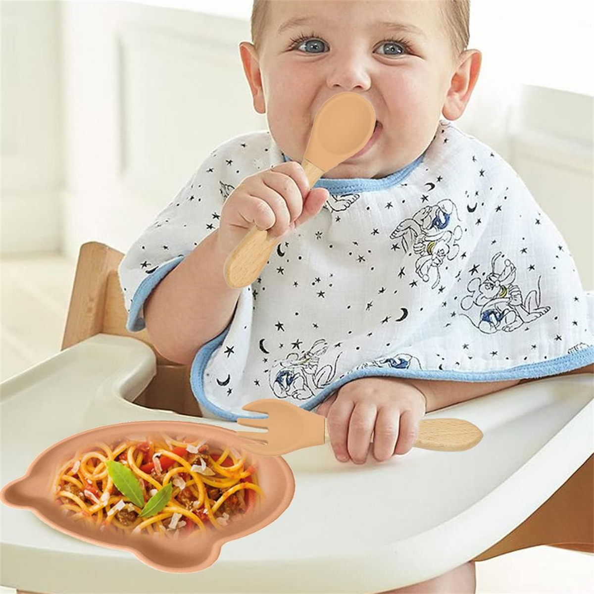 Baby im Hochstuhl, isst mit Löffel. Teller mit Pasta in der Nähe. Trägt Lätzchen mit Cartoonfiguren. Fröhlicher Ausdruck.