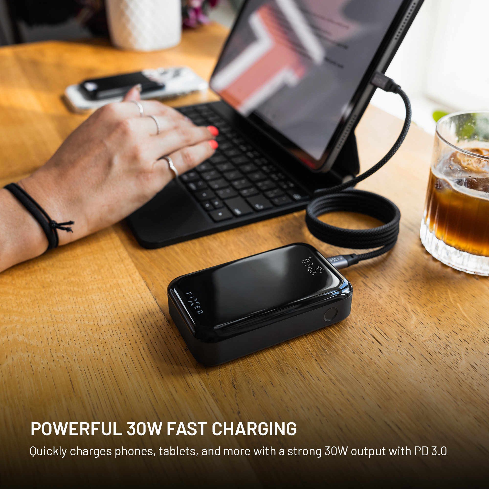 Eine schwarze Powerbank, die ein Tablet auflädt. Eine Hand tippt auf einer Tastatur. Im Hintergrund ein Glas Eiskaffee.