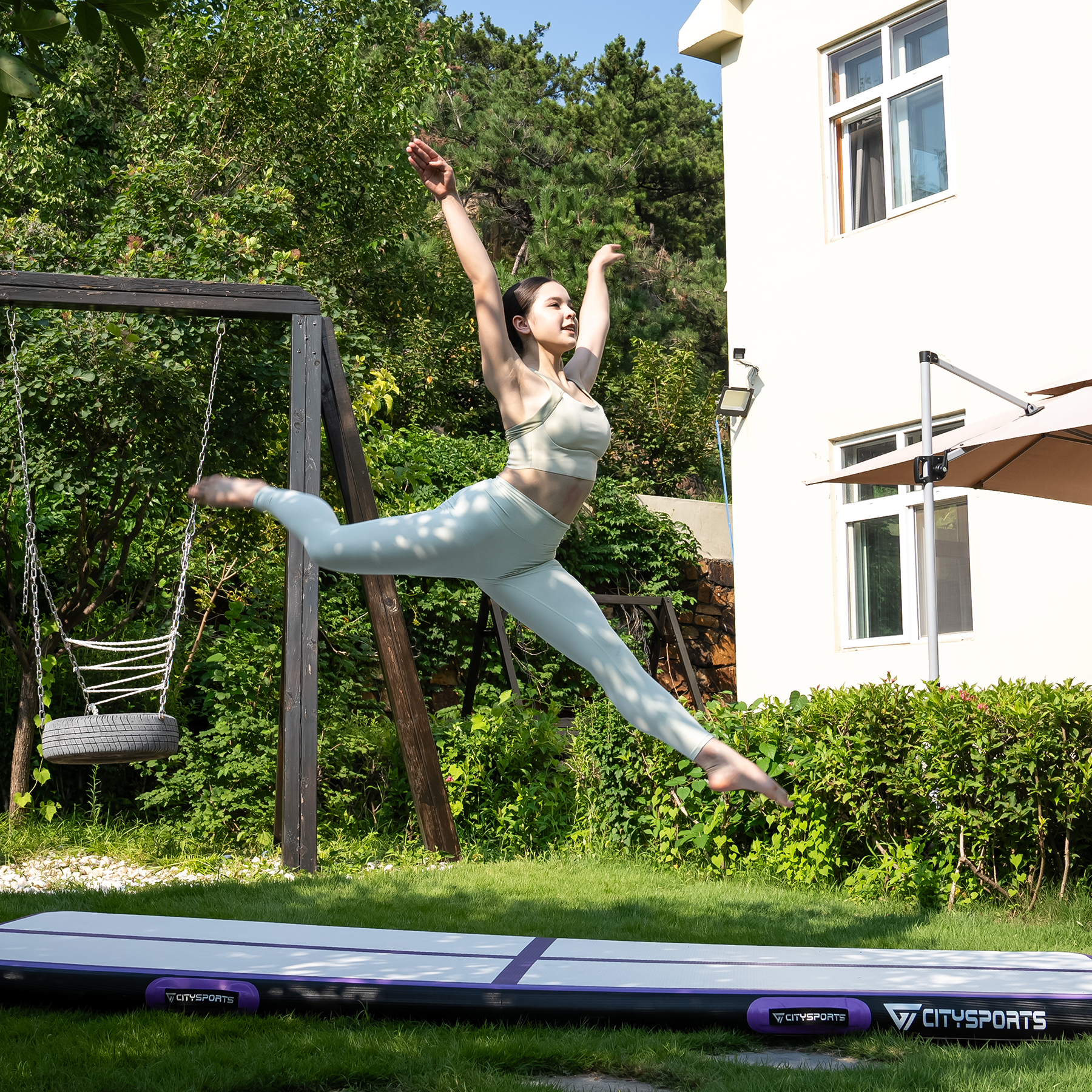 A woman jumps in the air on a mat. She wears athletic clothing. A swing set and a house are in the background.