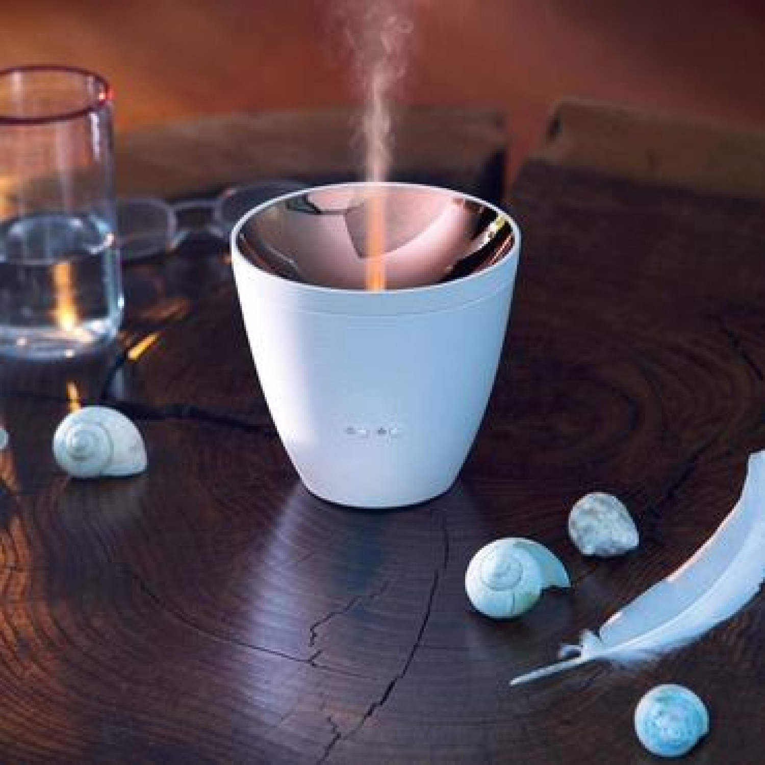 Ein weißer Aromadiffusor mit Dampf auf einem Holztisch. Muscheln, eine Feder und ein Glas Wasser befinden sich ebenfalls auf dem Tisch.