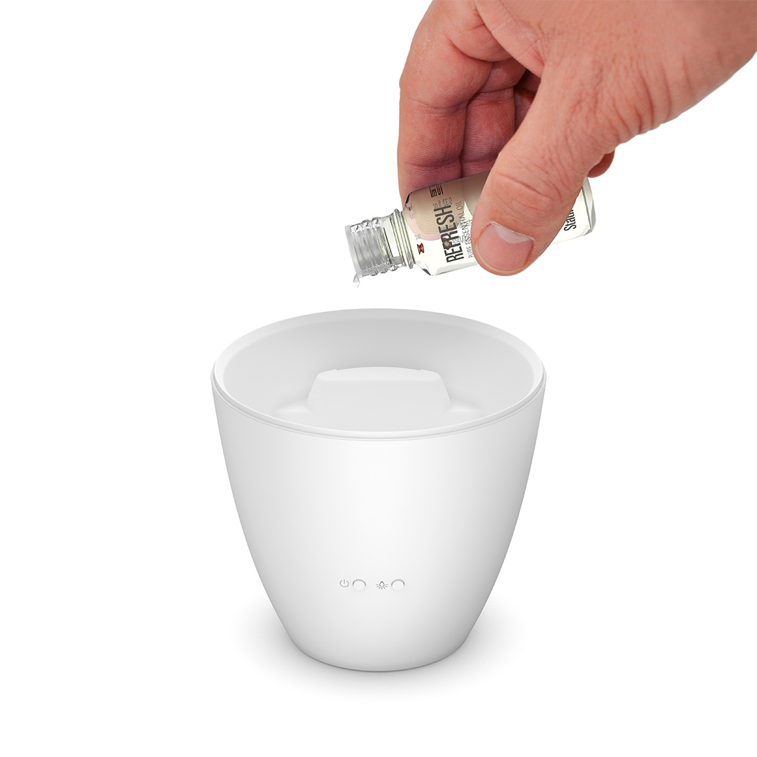 Eine Hand gießt Flüssigkeit aus einer kleinen Flasche mit der Aufschrift 'Refresh' in einen weißen Diffusor.