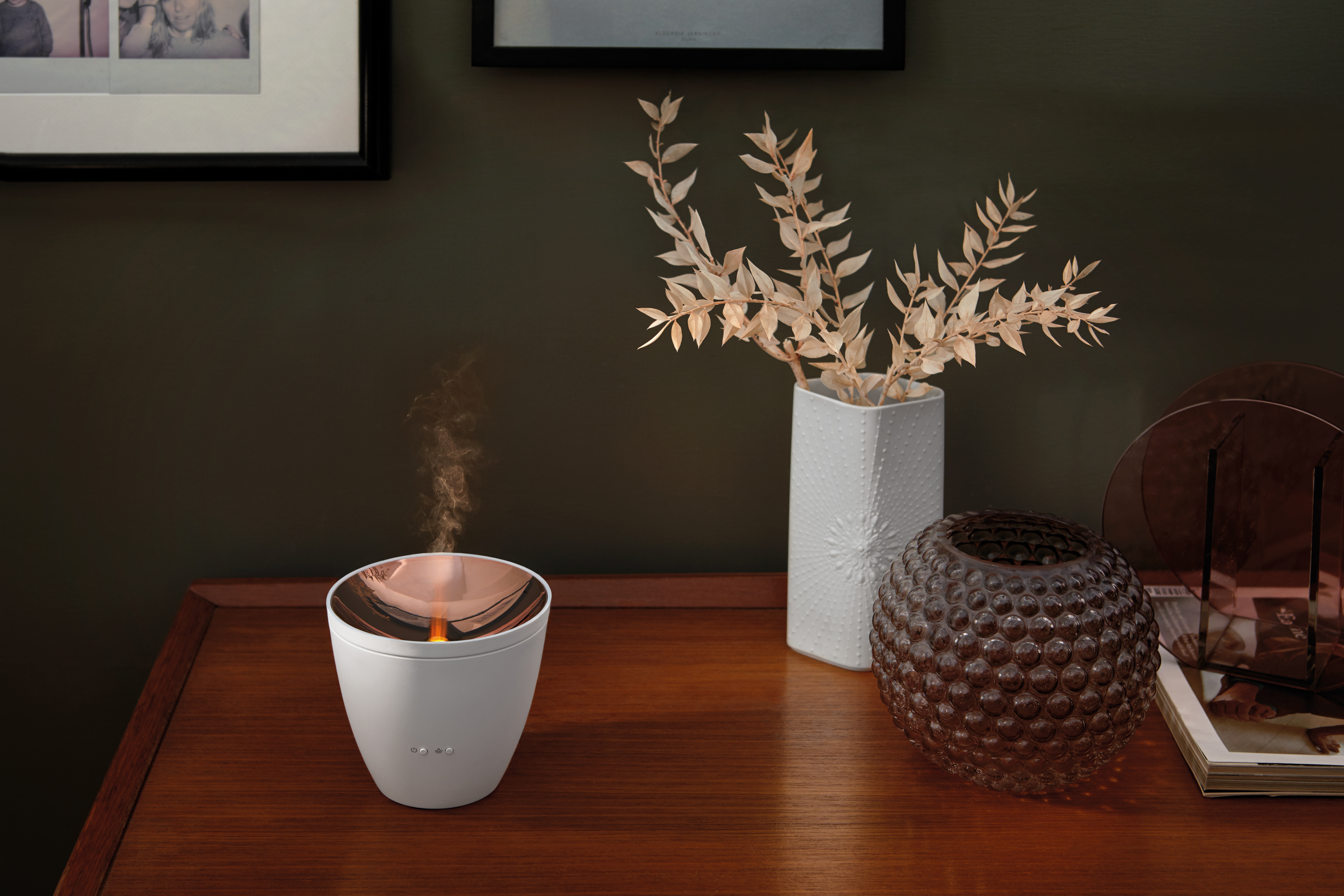Ein weißer Diffusor mit Dampf, eine Vase mit Zweigen und eine braune Glaskugel auf einem Holztisch.
