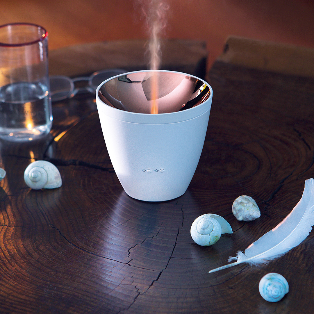 Ein weißer Diffusor mit Dampf, Muscheln, einer Feder und einem Glas Wasser auf einem Holztisch.