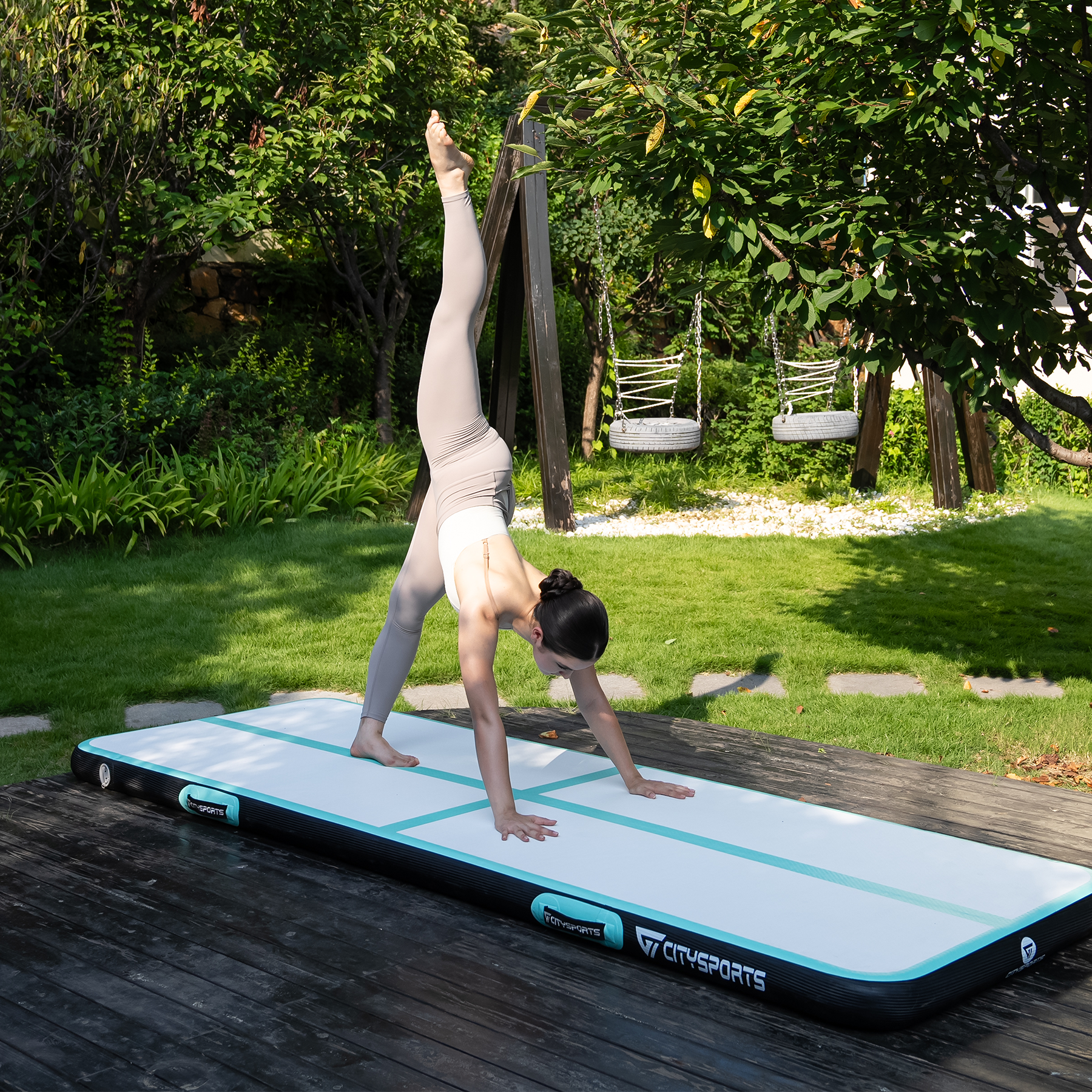 Kobieta w staniu na rękach na macie powietrznej na zewnątrz, z huśtawką w tle. Zielona trawa i drzewa otaczają.