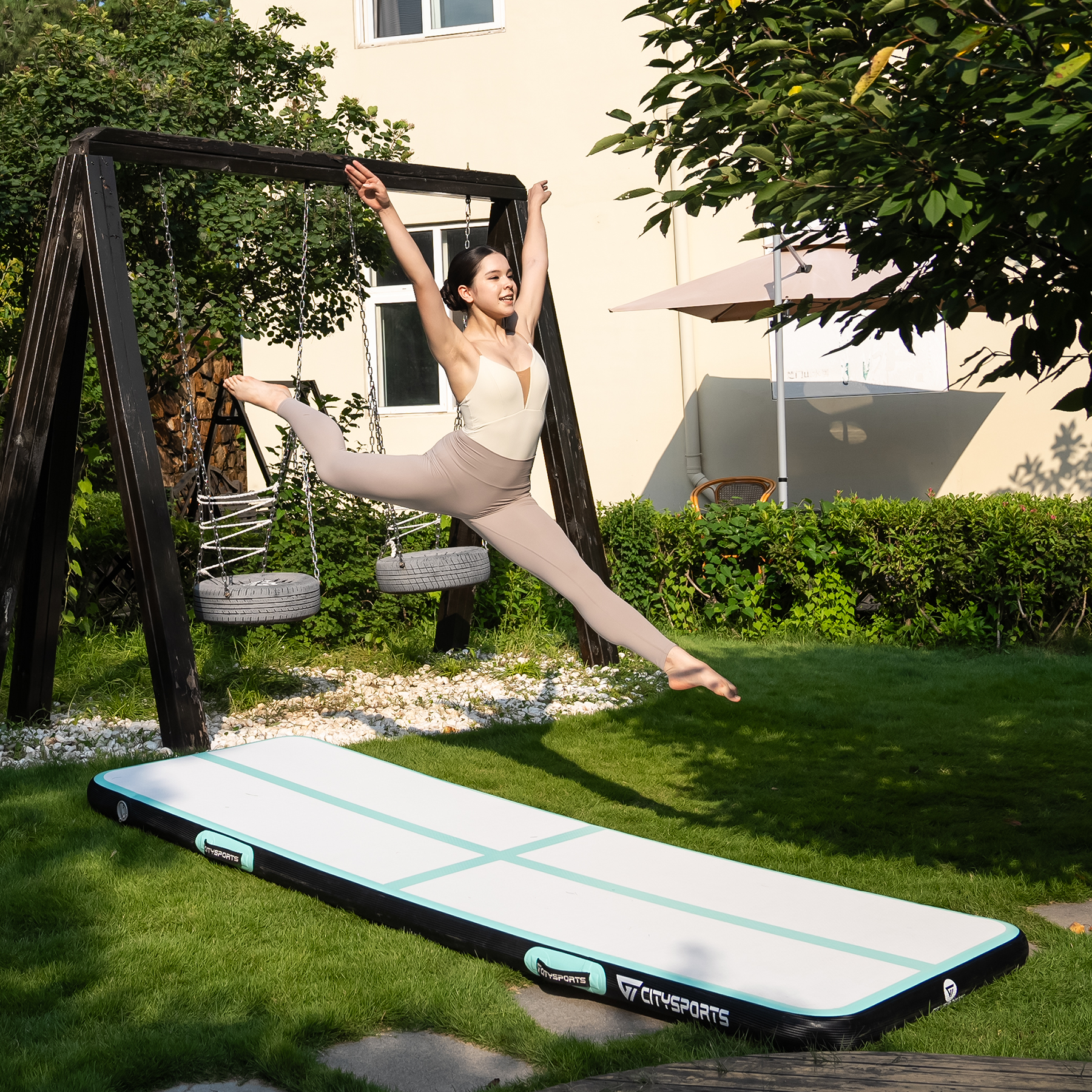 Kobieta wykonuje szpagat w powietrzu nad torem powietrznym w ogrodzie. W tle huśtawka i budynek.