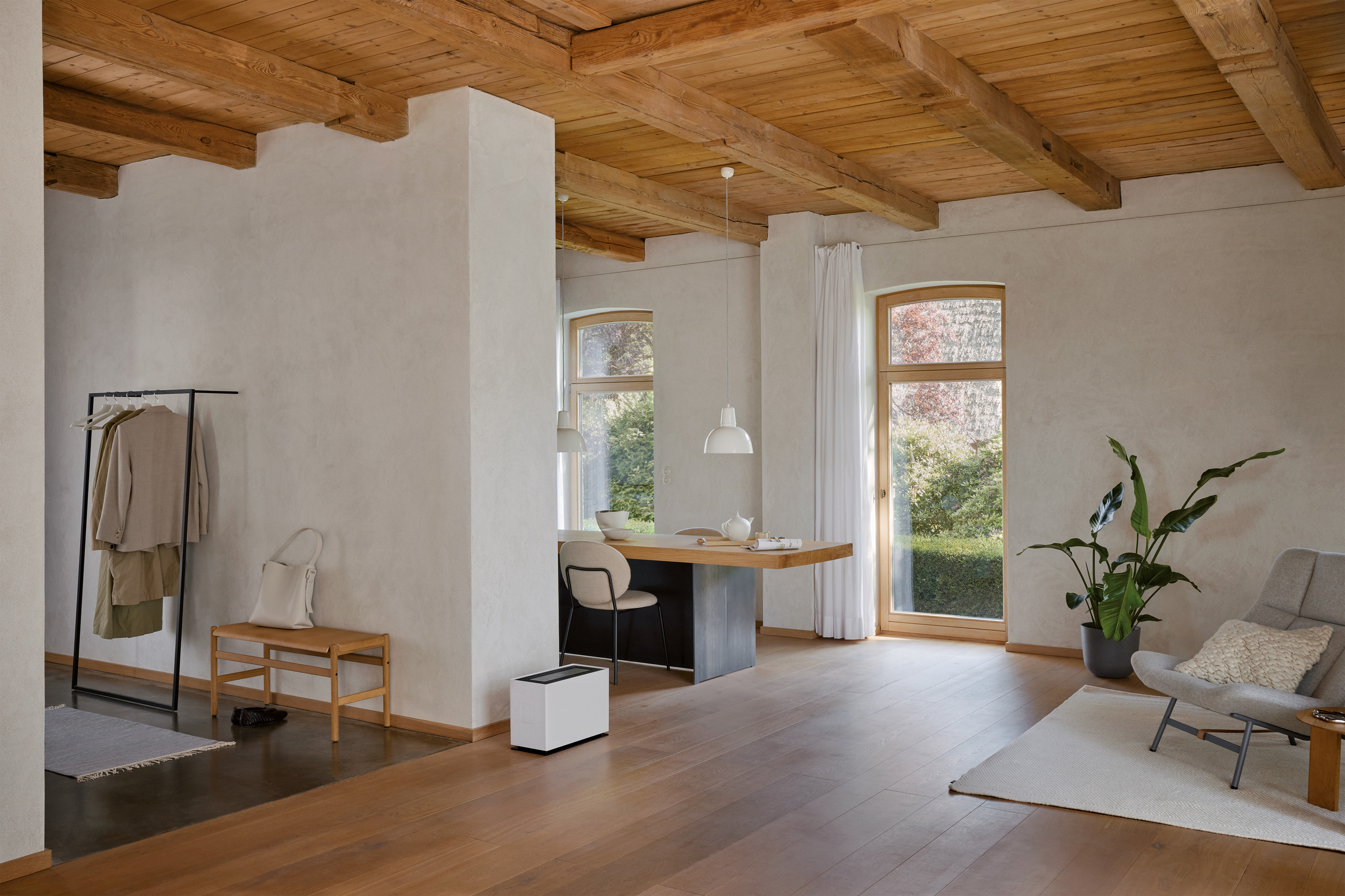 Innenraum mit Holzdecke, weißen Wänden und Holzboden. Enthält Schreibtisch, Stuhl, Garderobe und Pflanze. Große Fenster bieten Tageslicht.