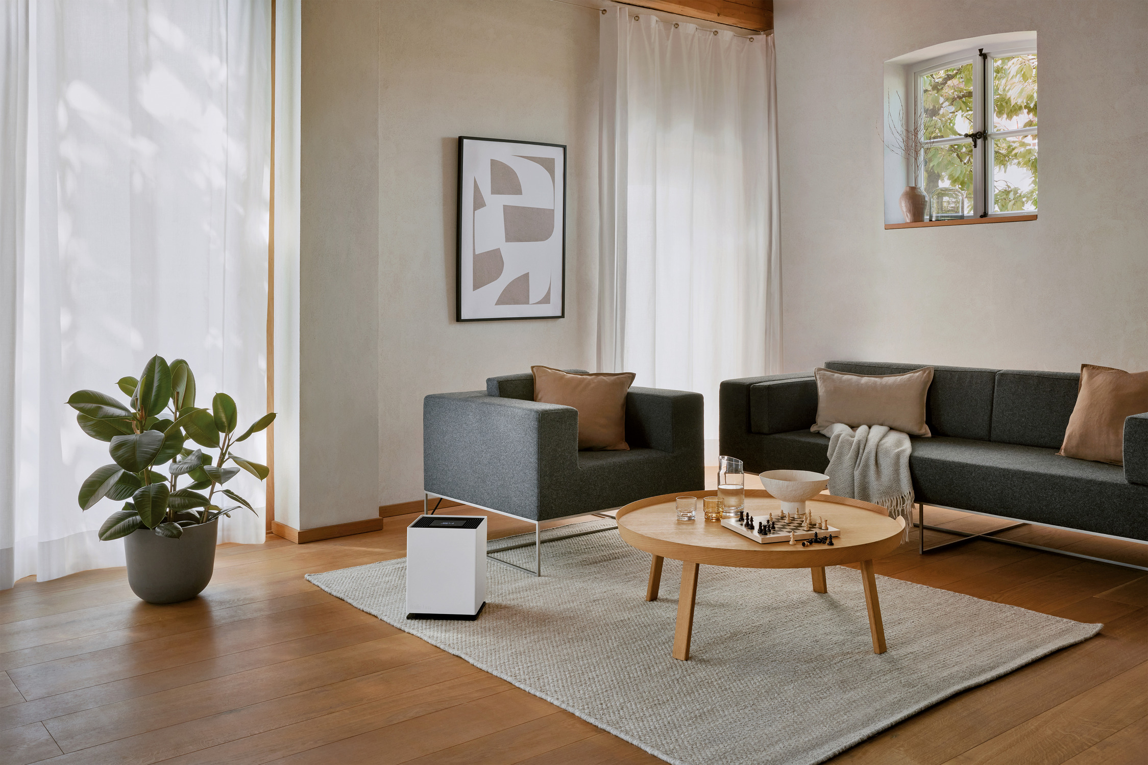 Wohnzimmer mit grauem Sofa und Sesseln, Holztisch, großer Pflanze und Fenster. Ein gerahmtes abstraktes Kunstwerk an der Wand.