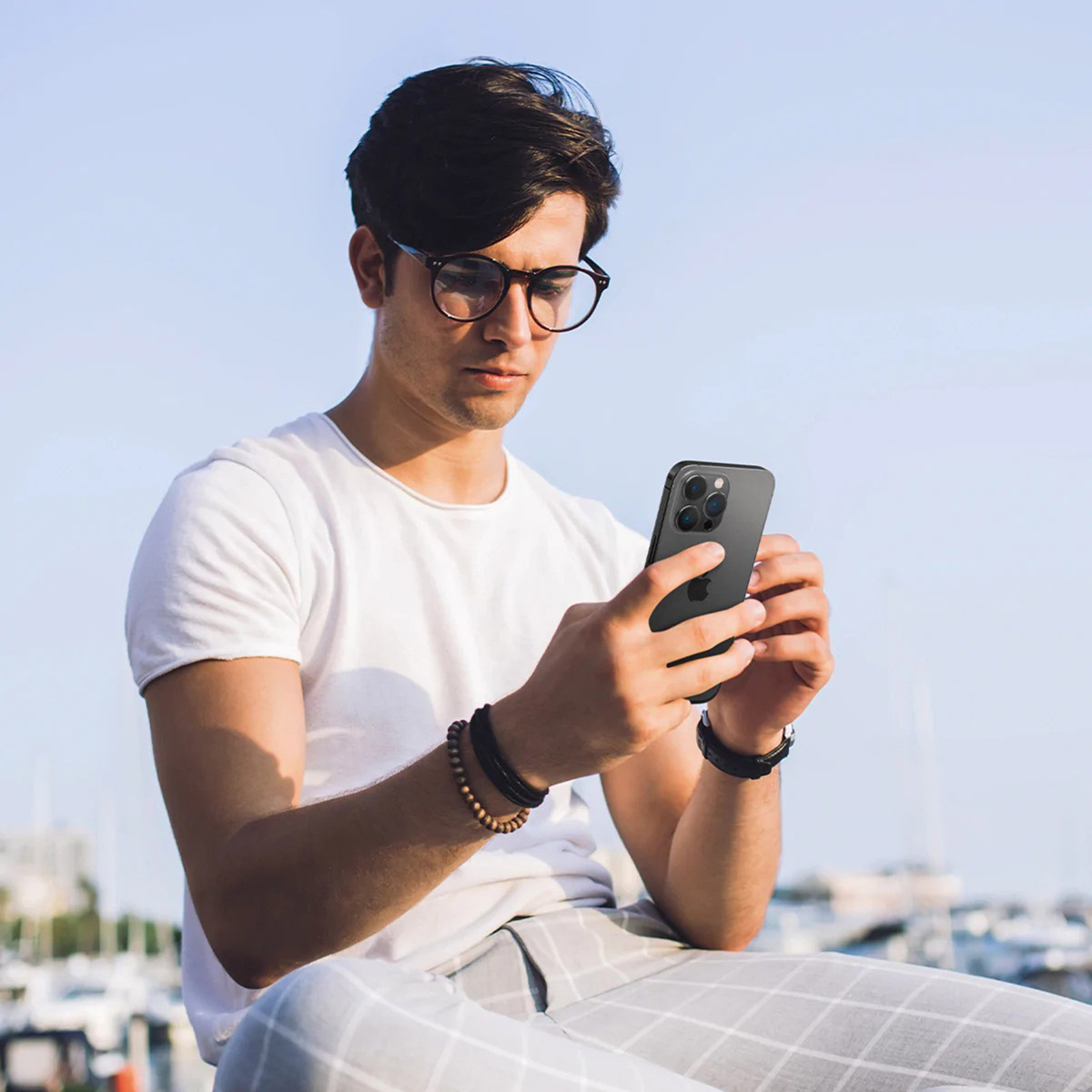 Un homme avec des lunettes tient un téléphone noir. Il porte une chemise blanche, des bracelets et une montre. Le fond a des bateaux.