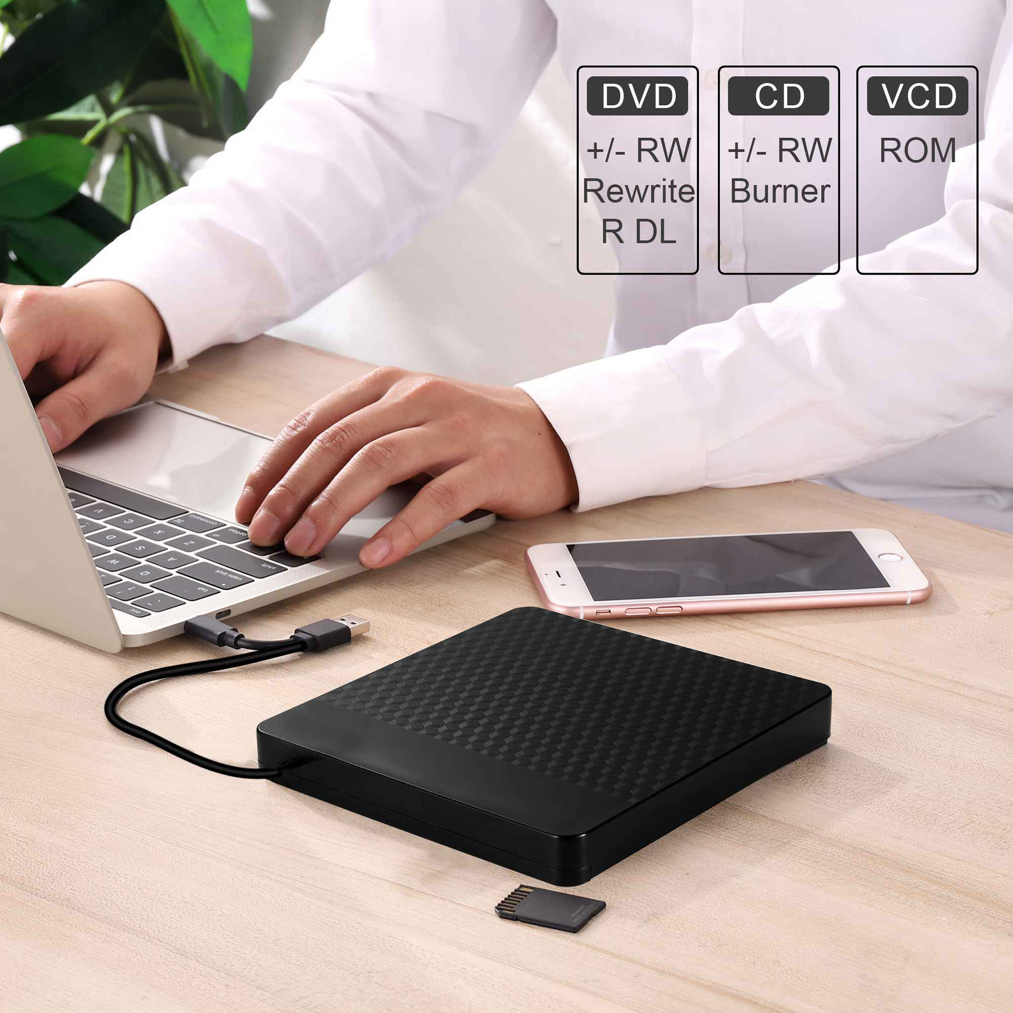 Eine Person arbeitet an einem Laptop. Auf dem Tisch befinden sich ein schwarzes externes DVD-Laufwerk, ein Telefon und eine Speicherkarte.