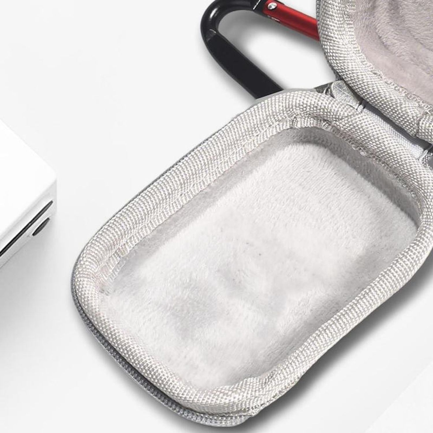 Custodia grigia aperta con interno bianco e soffice, moschettone nero e rosso. Superficie bianca sullo sfondo, parte di un dispositivo bianco.
