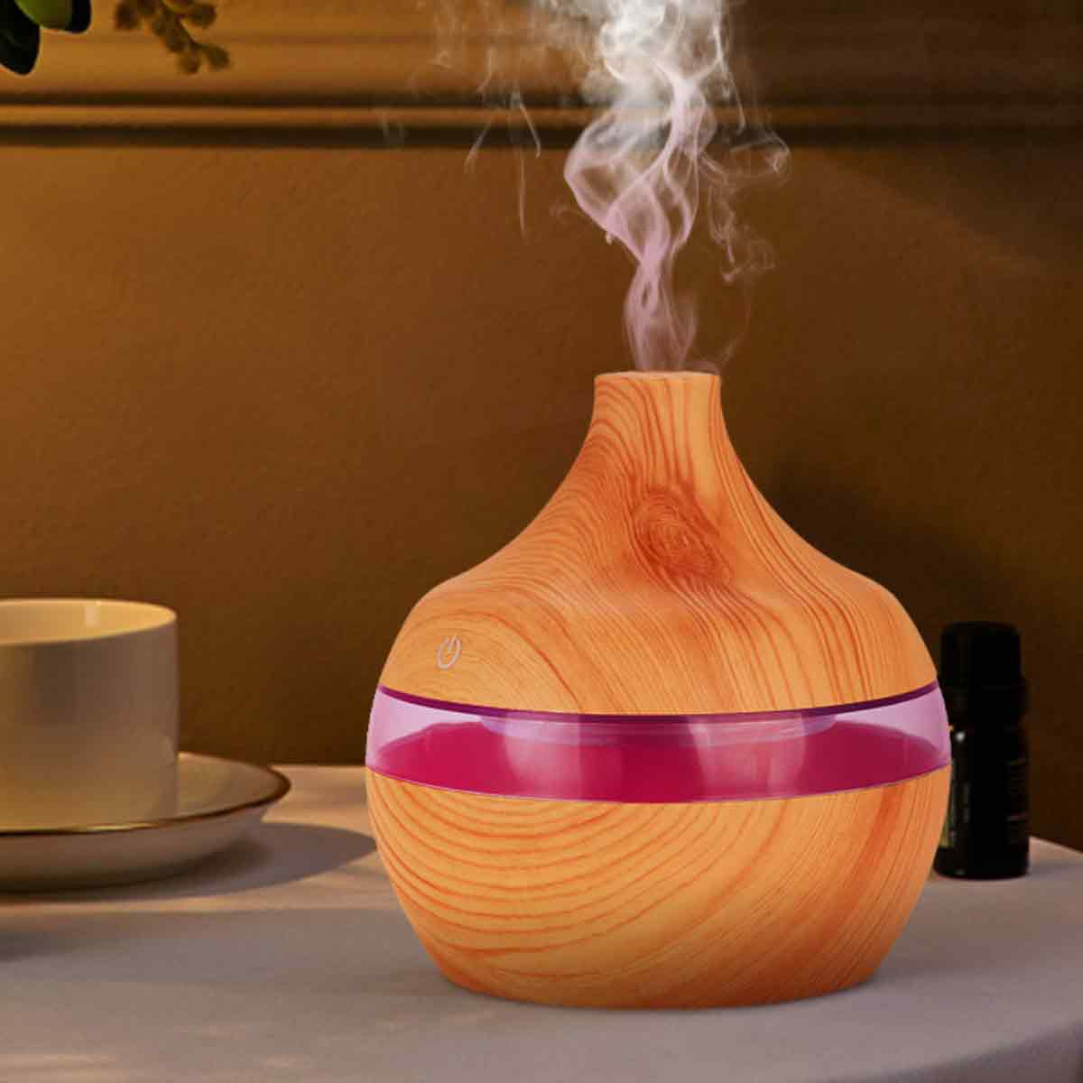 Ein hölzerner Aromadiffusor mit rosa und klaren Akzenten gibt weißen Dampf ab. Eine Tasse und eine kleine schwarze Flasche stehen auf dem Tisch.