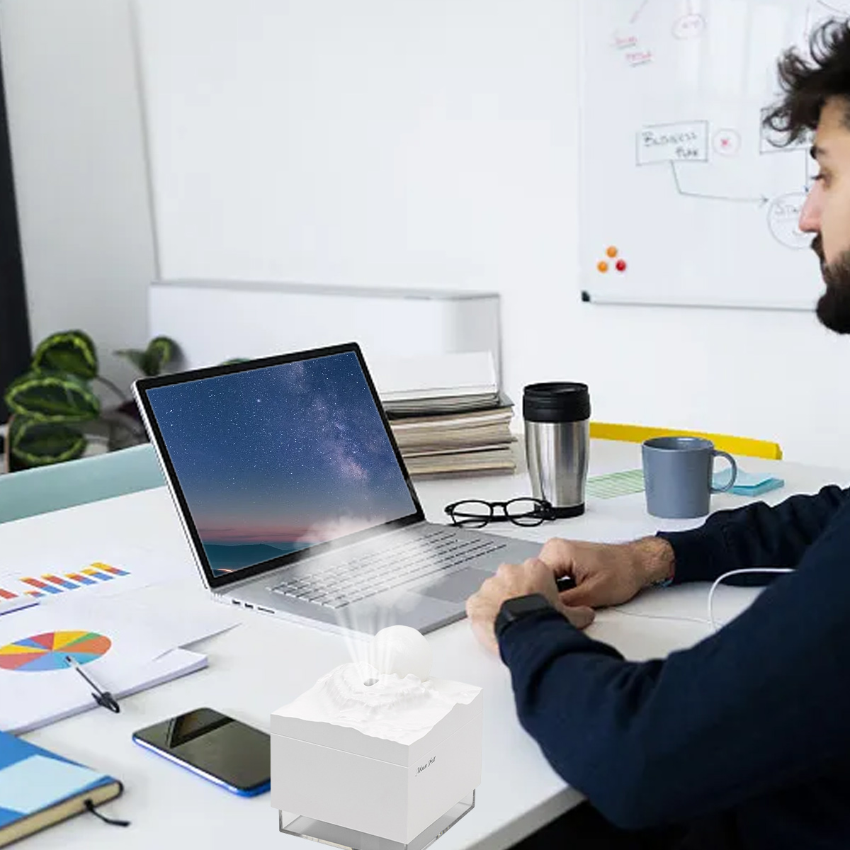 Ein Mann sitzt an einem Schreibtisch mit einem Laptop. Auf dem Schreibtisch befinden sich ein Vulkanmodell, ein Telefon, eine Brille, eine Tasse und Papiere.