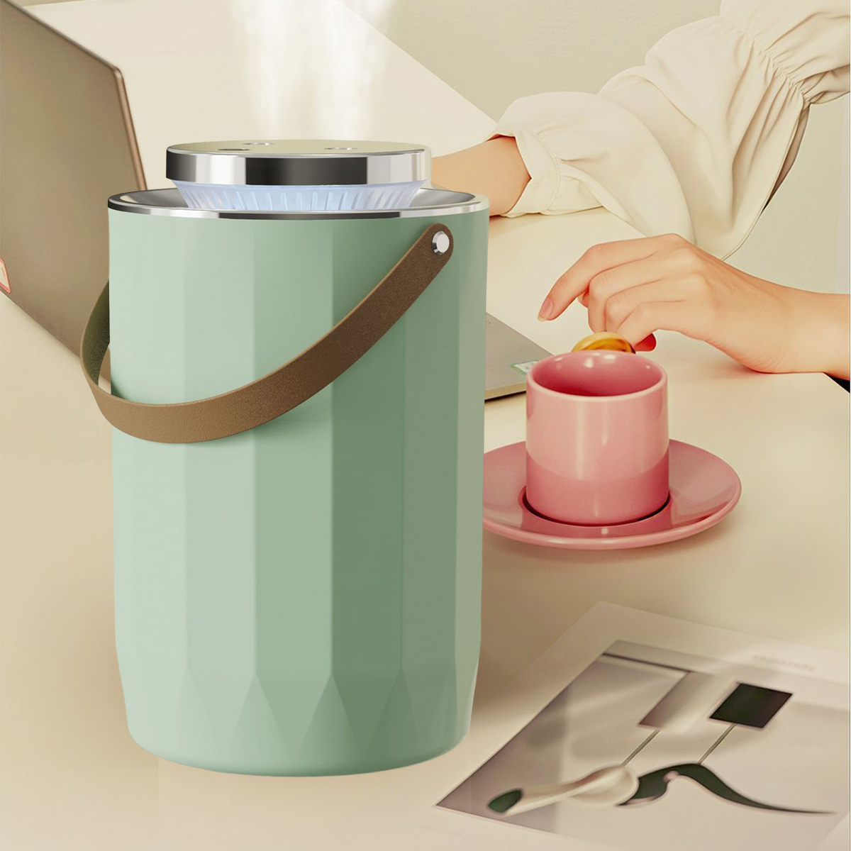 Ein mintgrüner Luftbefeuchter mit braunem Griff steht auf einem Schreibtisch. Eine rosa Tasse und Untertasse sind in der Nähe. Eine Person greift nach der Tasse.