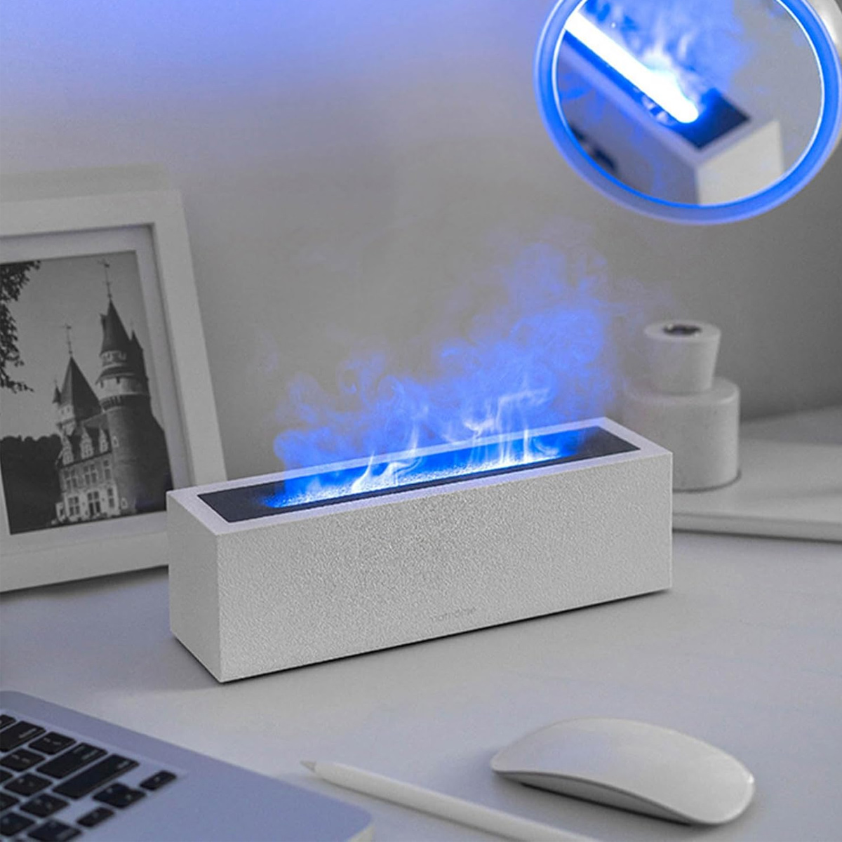 Ein weißer, rechteckiger Luftbefeuchter stößt blauen Dampf aus, der Flammen nachahmt. Ein Bilderrahmen, eine Maus, ein Bleistift und ein Laptop sind auf dem Tisch.