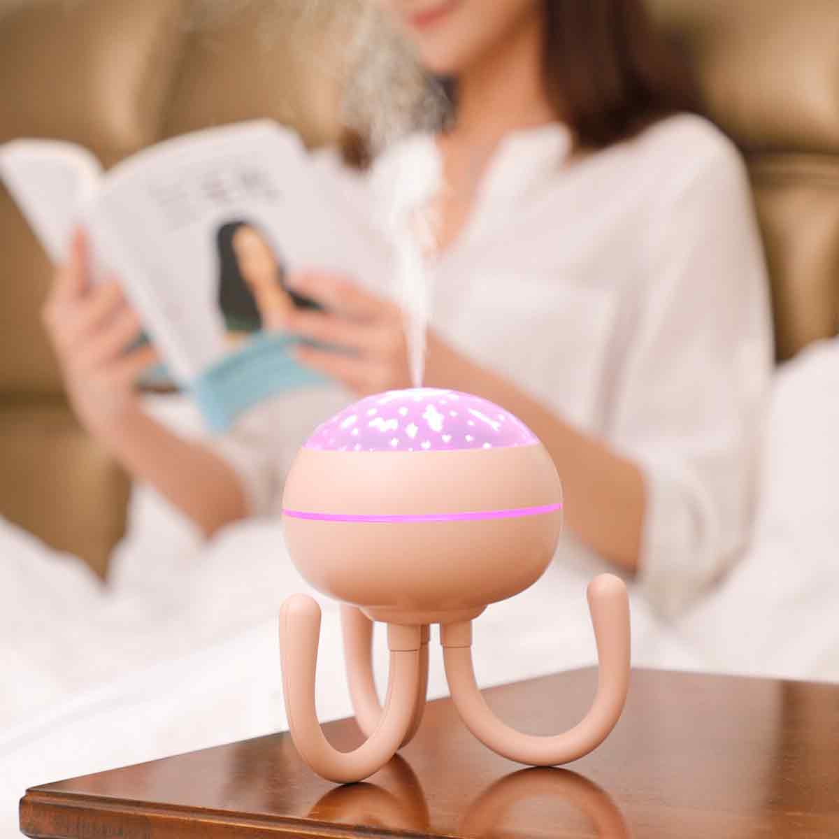 Ein rosa Luftbefeuchter mit leuchtenden Sternen steht auf einem Tisch. Im Hintergrund liest eine Frau in weißer Kleidung.