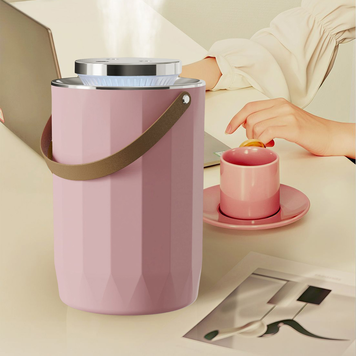Ein rosa zylindrisches Gerät, eine Hand, die eine Tasse hält, und ein Laptop befinden sich auf einer hellen Oberfläche. Rechts befinden sich eine Tasse und eine Untertasse.
