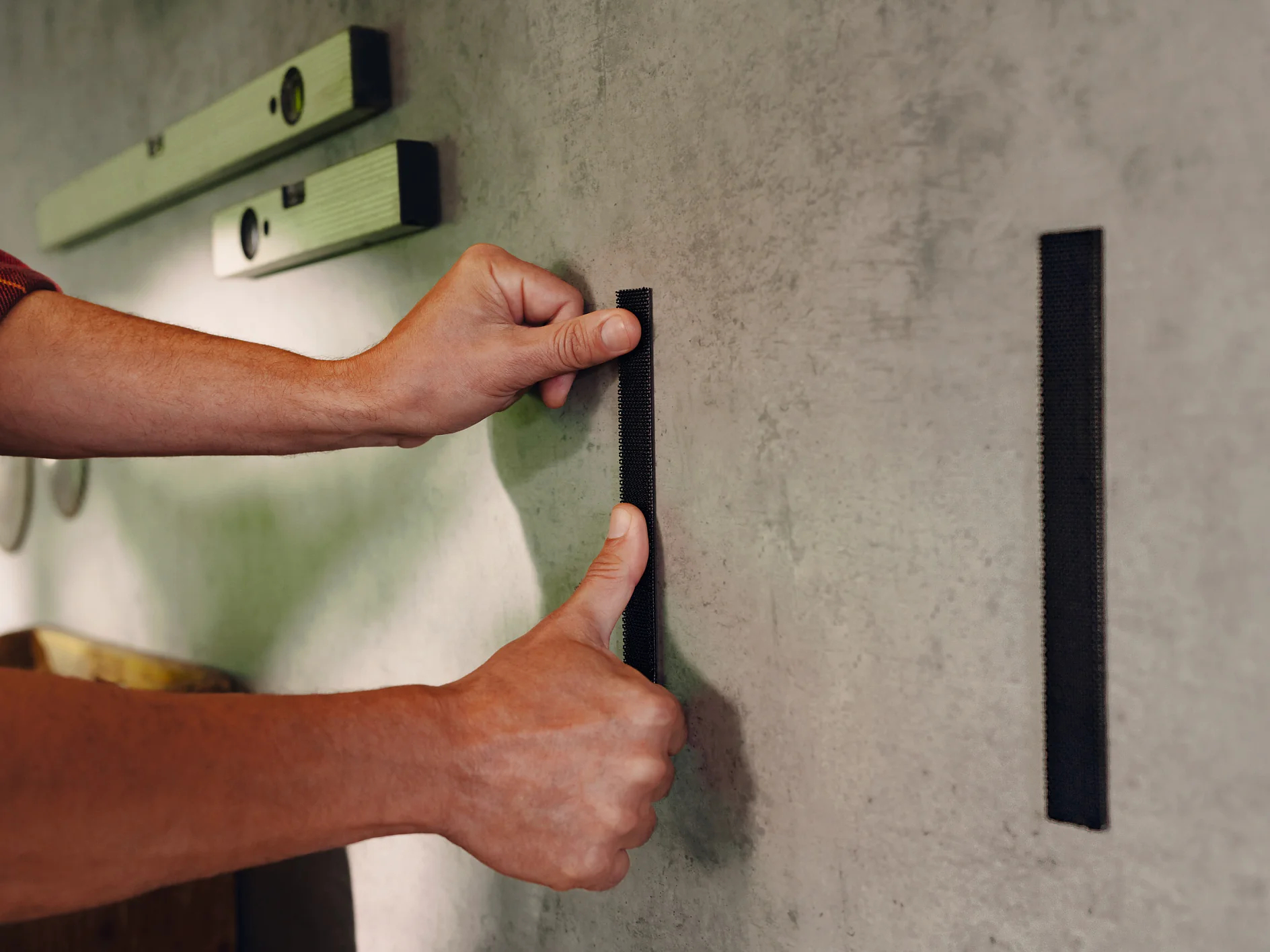 Hände einer Person befestigen einen schwarzen Streifen an einer grauen Wand. Zwei Wasserwaagen sind darüber montiert. Heimwerken.
