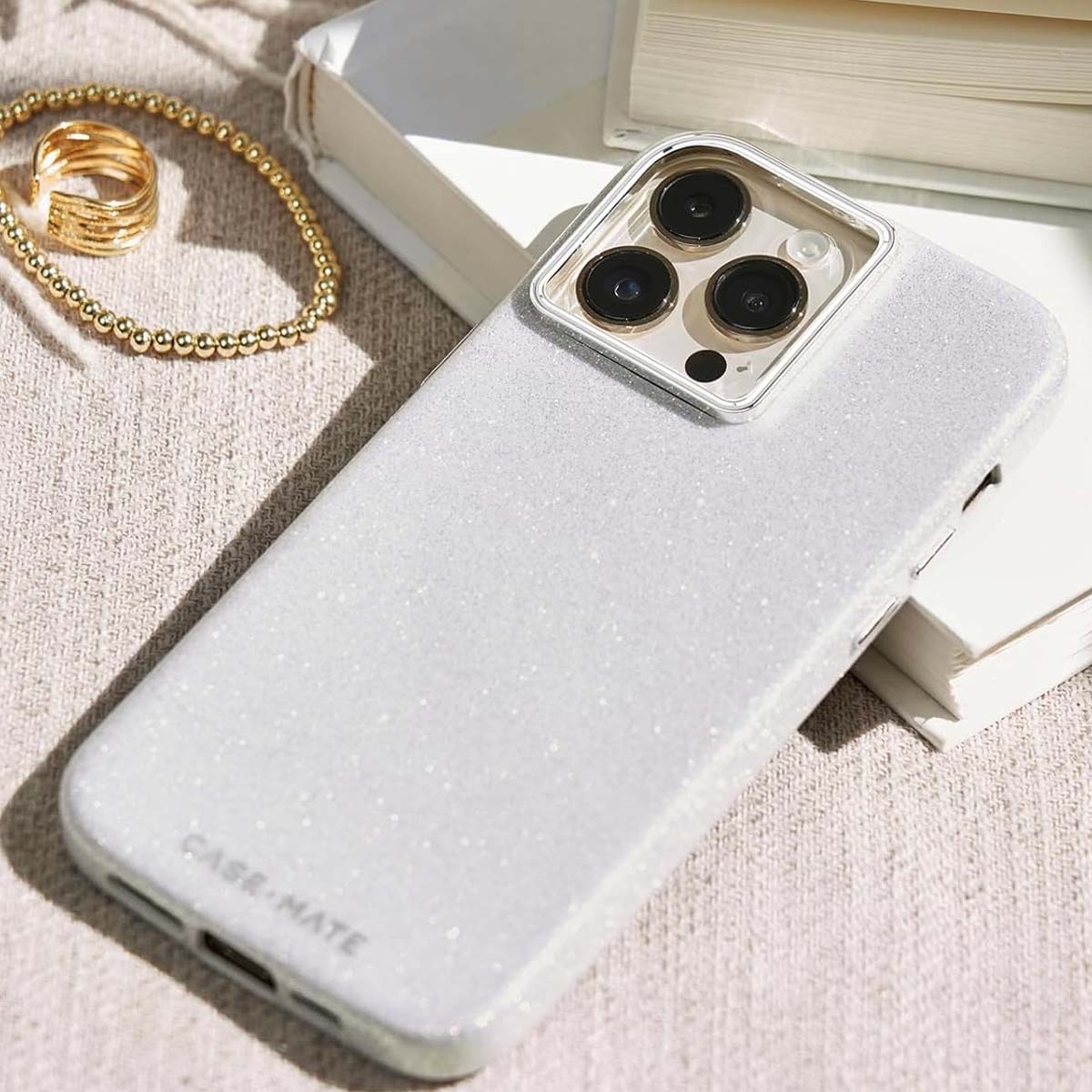 Una custodia bianca glitterata per iPhone di Case-Mate è su un libro. Un anello e un braccialetto dorati sono su un tessuto beige.