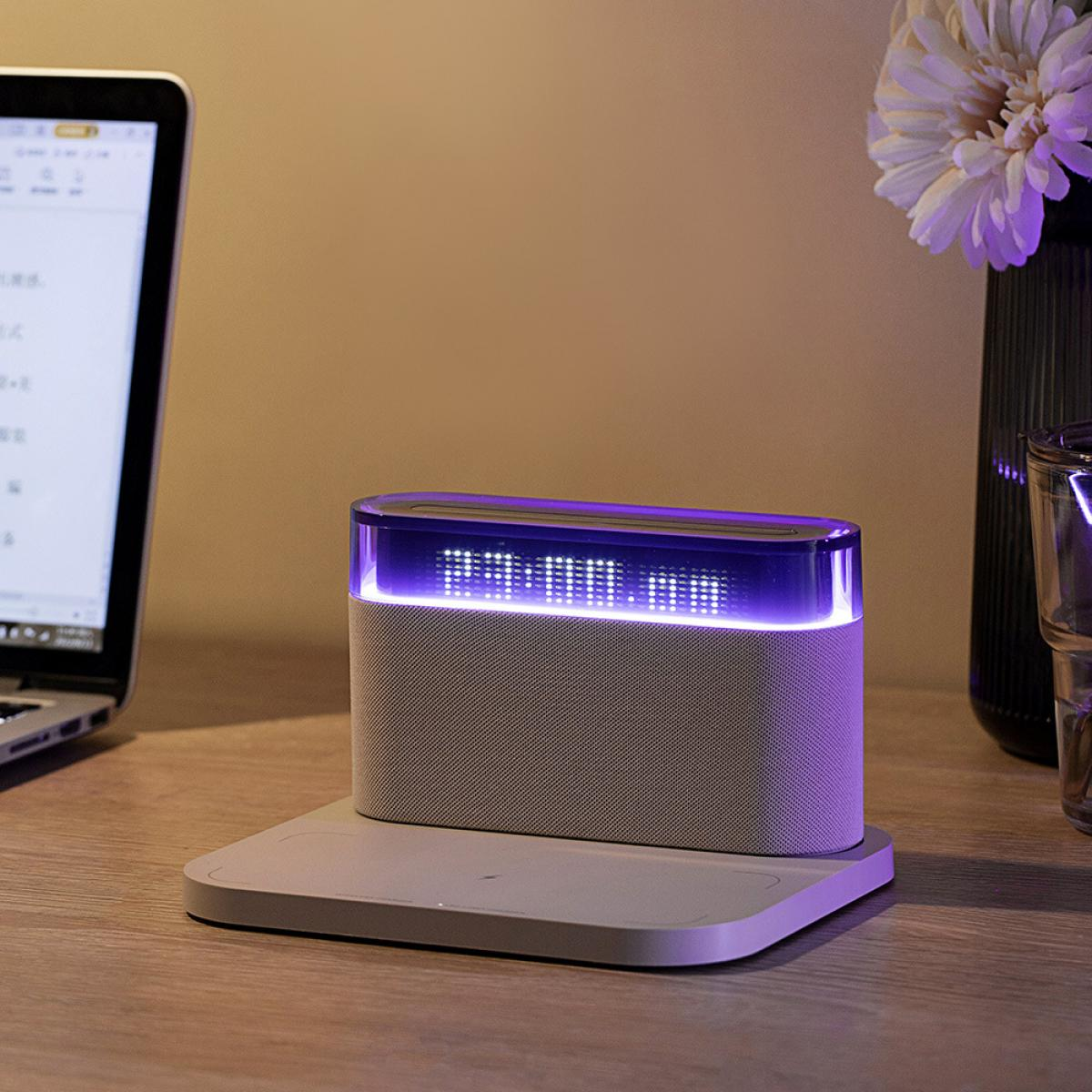 Eine Digitaluhr zeigt die Uhrzeit an. Sie steht auf einer grauen Basis auf einem Holztisch, mit einem Laptop und einer Blumenvase im Hintergrund.