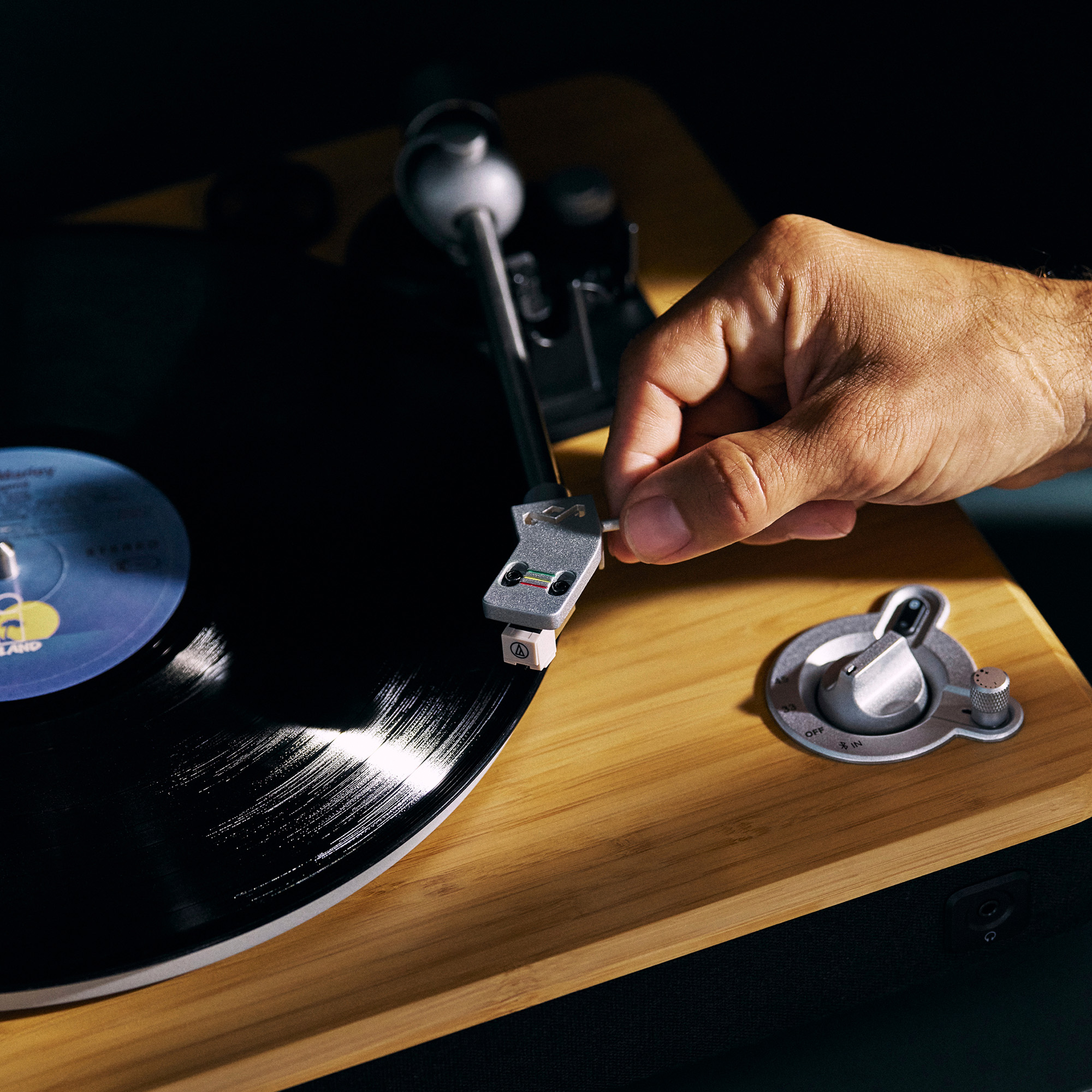 Eine Hand senkt die Nadel auf eine Vinyl-Schallplatte auf einem hölzernen Plattenspieler ab. Die Platte ist schwarz mit blauem Etikett.