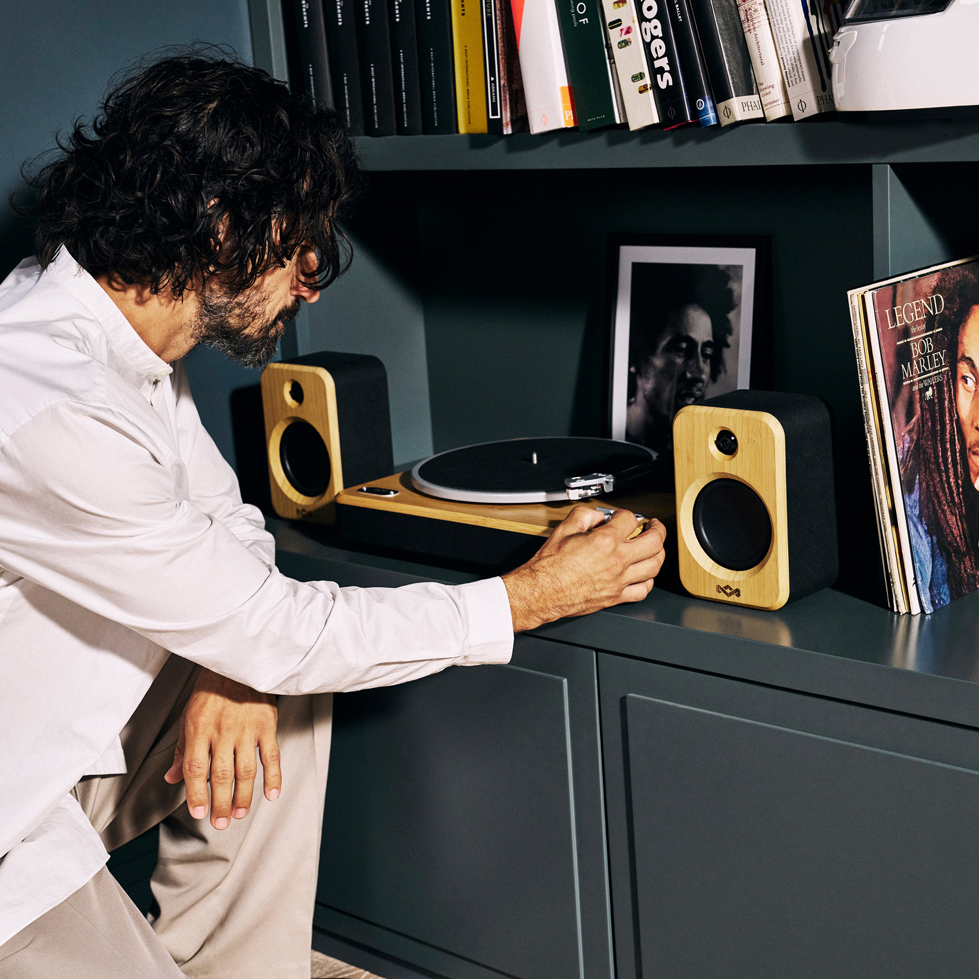 Ein Mann justiert einen Plattenspieler. Lautsprecher, Bob Marley-Album, Bücher und ein Foto stehen auf einem Regal.
