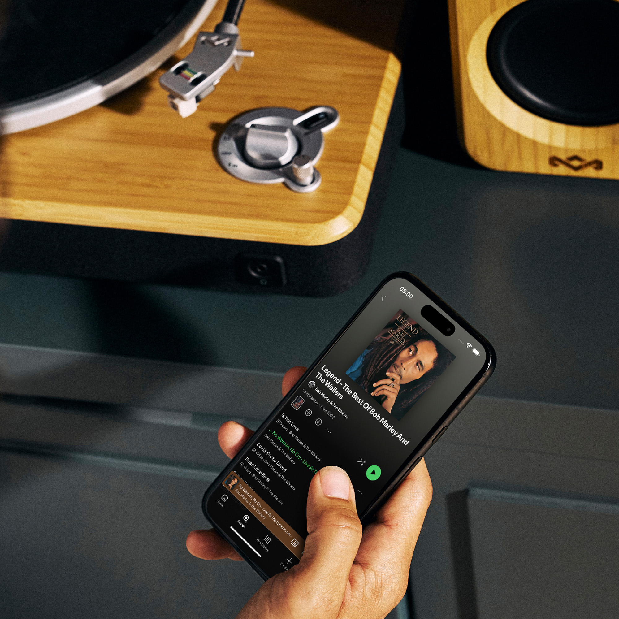 Eine Hand hält ein Telefon mit einem Bob Marley-Album. Im Hintergrund ein Plattenspieler und ein Lautsprecher.