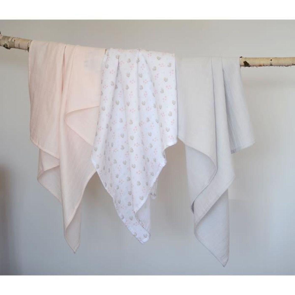 Drei Babydecken hängen an einem Ast. Eine rosa, eine mit rosa Blumen gemustert und eine graue. Der Hintergrund ist weiß.