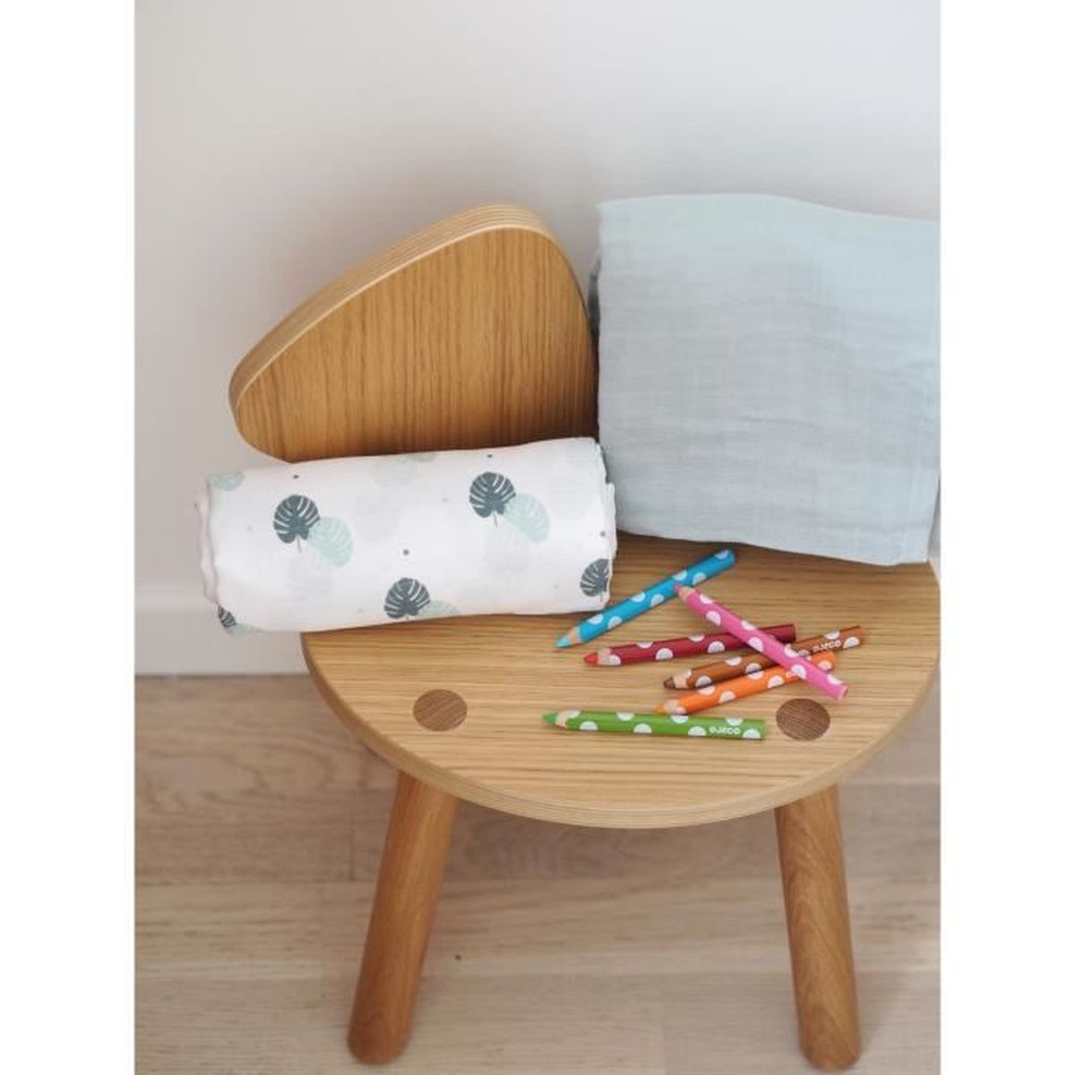 Ein Holzschemel mit einer gerollten Decke, einem blauen Tuch und Buntstiften. Die Decke hat Blättermuster. Buntstifte in verschiedenen Farben sind verstreut.