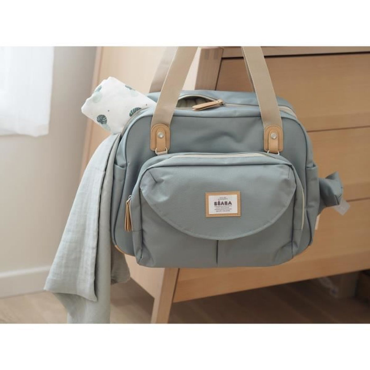 Eine hellblaue Babytasche mit beigefarbenen Riemen und einer passenden Vordertasche. Eine Decke mit Blättermuster hängt an der Seite.