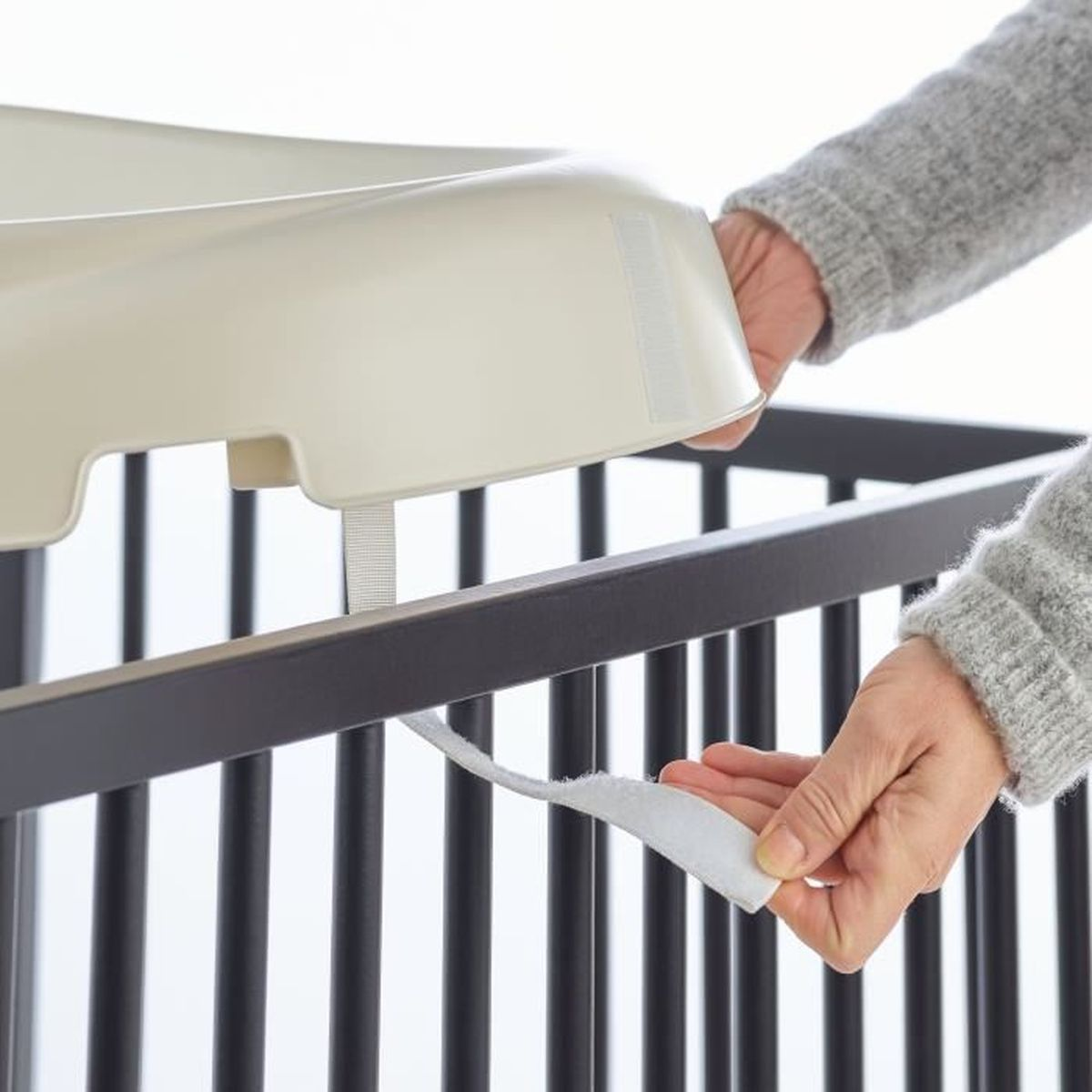 Hände befestigen eine hellfarbene Abdeckung an einem dunklen Kinderbett. Die Abdeckung hat einen Klettverschluss. Die Person trägt einen grauen Pullover.