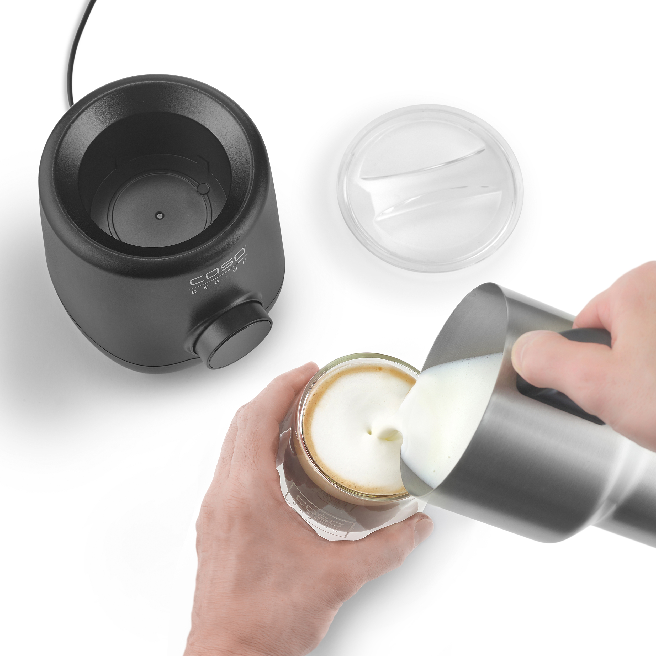 Eine Person gießt Milchschaum in ein Glas Kaffee. Ein schwarzer Aufschäumer und ein Deckel sind sichtbar.