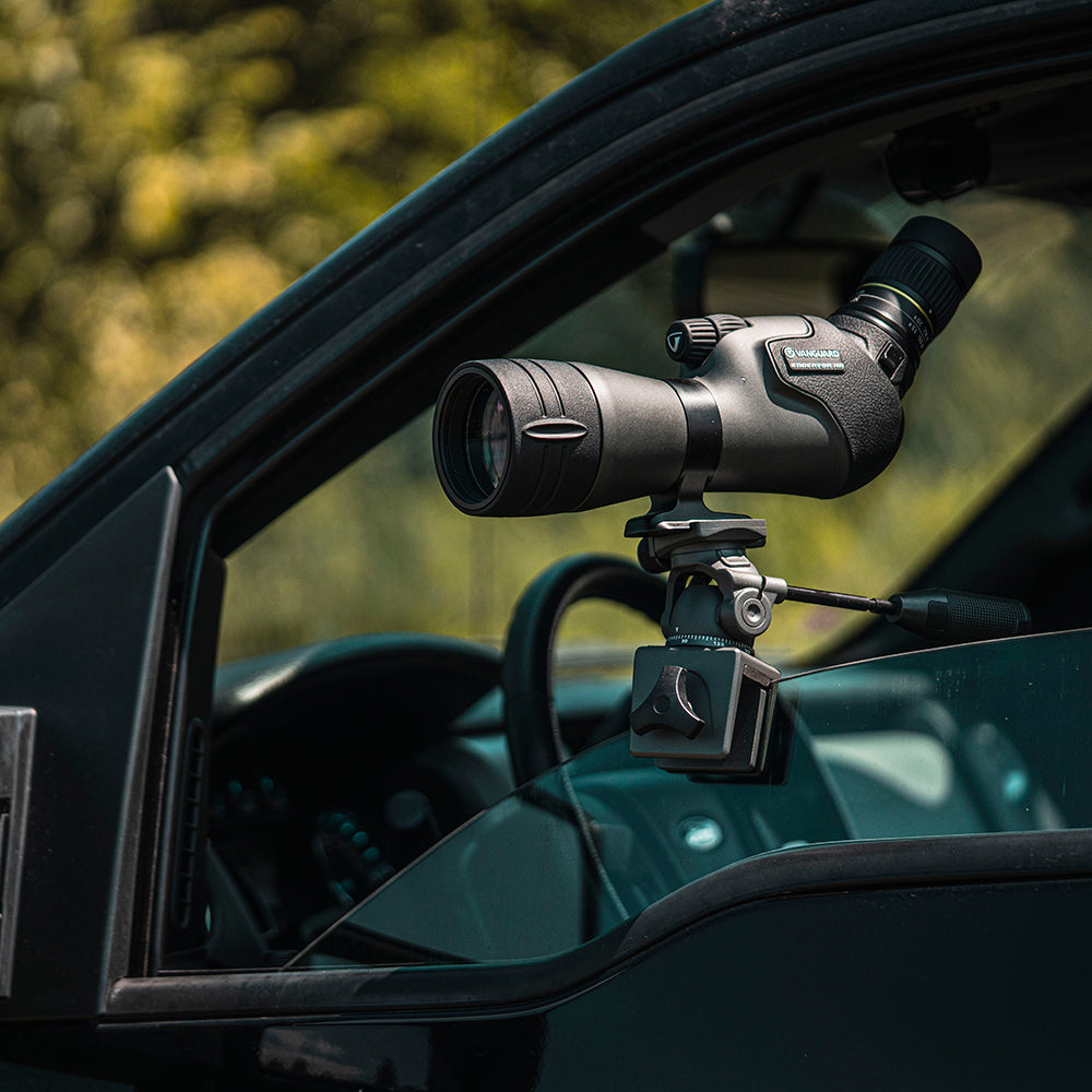 Un telescopio terrestre negro montado en un trípode está dentro de un coche. El telescopio apunta hacia un fondo verde borroso.