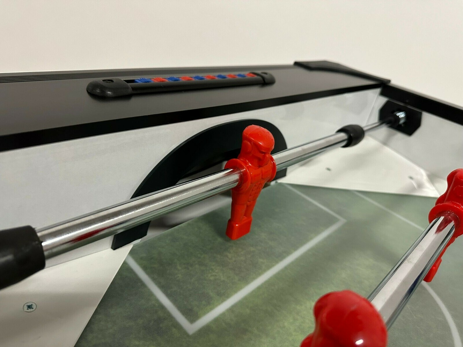 Close-up van een tafelvoetbaltafel. Rode spelersfiguren op metalen staven, groen veld, zwart-witte tafel met blauwe en rode scoremarkeringen.