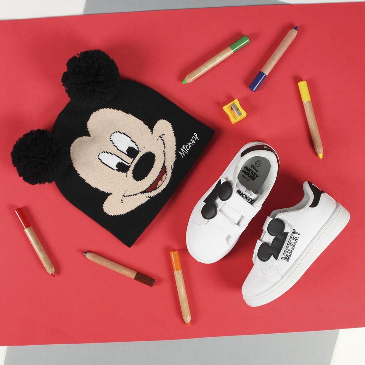 Un gorro negro de Mickey Mouse y zapatillas blancas con detalles de Mickey Mouse sobre una superficie roja. Lápices de colores dispersos.