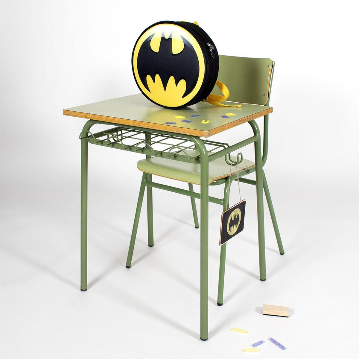 Escritorio escolar con una mochila de Batman. También hay una silla, un letrero de Batman y objetos pequeños. El fondo es blanco.