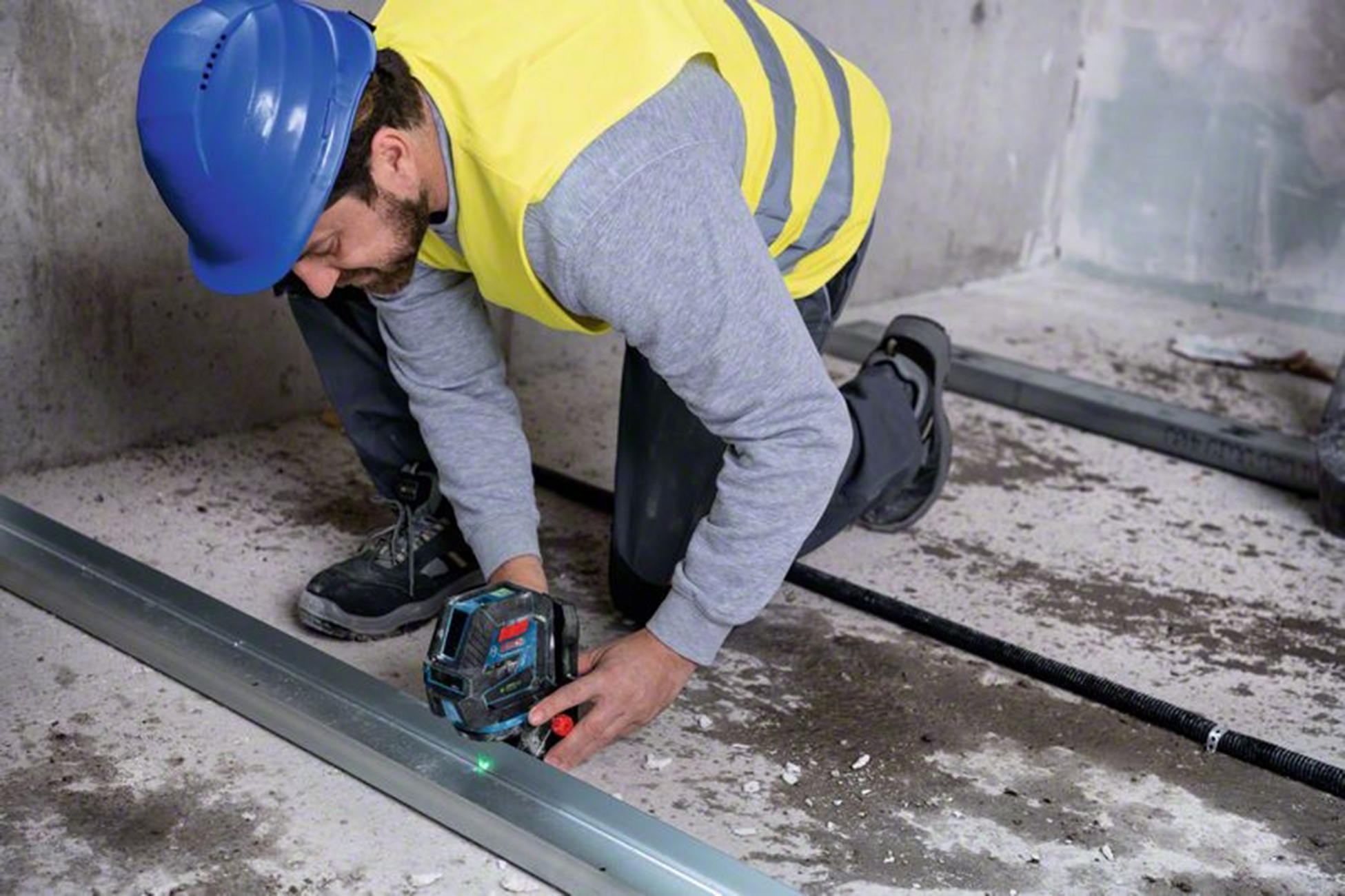 Bir işçi diz çökmüş, metal bir kirişte lazer seviyesi kullanıyor. Mavi bir kask ve sarı bir yelek giyiyor, beton bir odada.