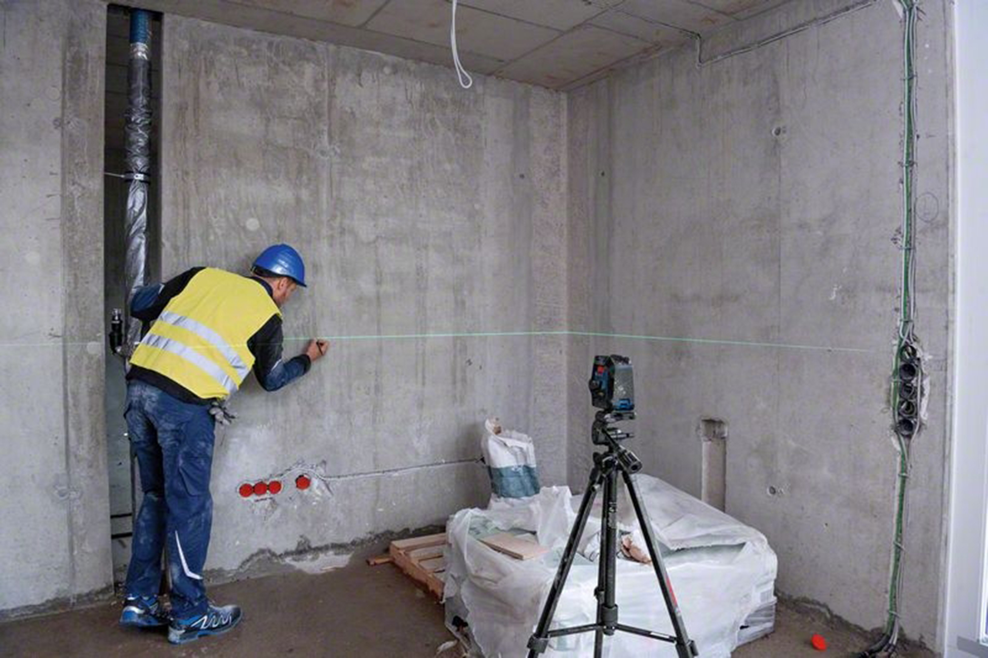 Sarı yelekli ve mavi kasklı bir inşaat işçisi, bir beton duvara bir çizgi çiziyor, bir tripod üzerinde lazer seviyesi ile.