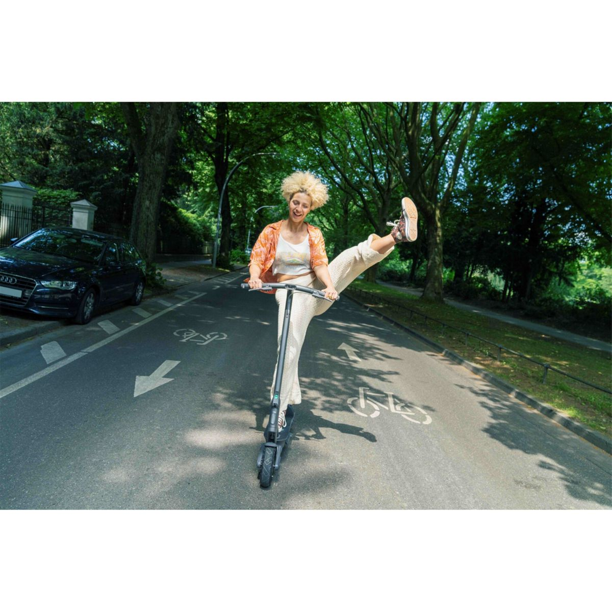 Eine Frau auf einem Elektroroller hebt ihr Bein. Sie befindet sich auf einer Straße mit Radwegmarkierungen, Bäumen im Hintergrund und einem geparkten Auto.