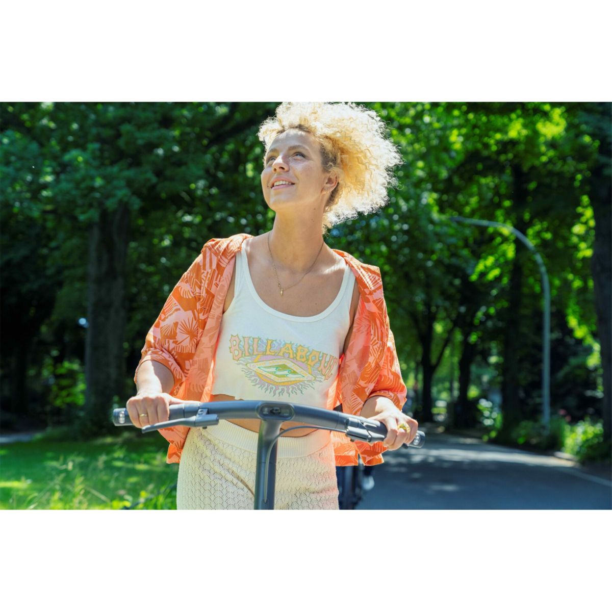 Frau auf Roller, blickt nach oben. Trägt orangefarbenes Oberteil, weißes Tanktop. Bäume im Hintergrund, sonniger Tag.