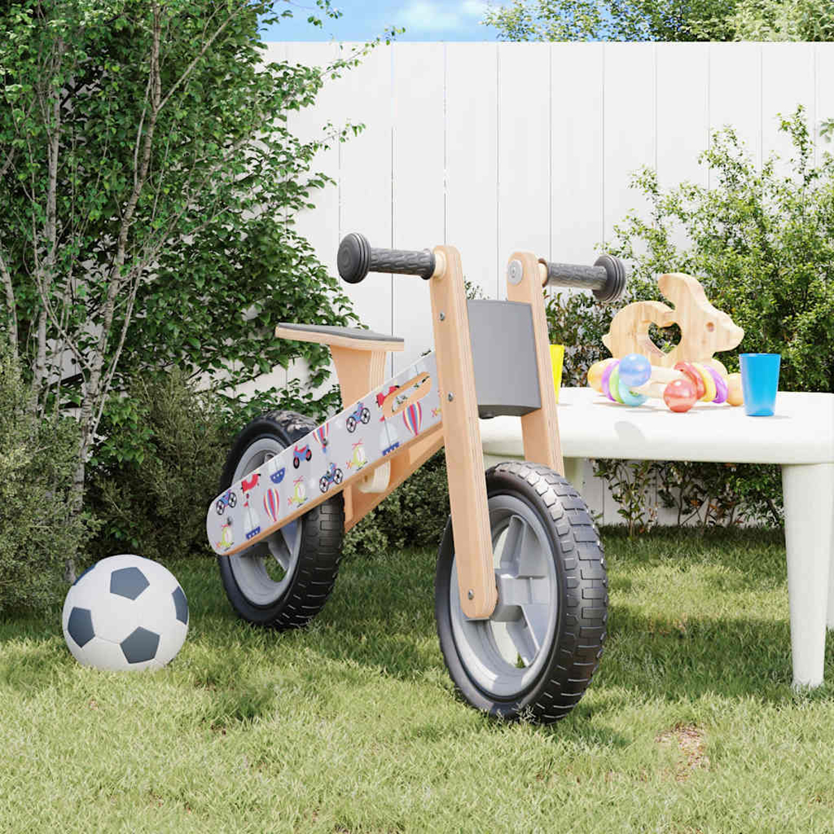 Ein hölzernes Laufrad steht auf Gras in der Nähe eines Fußballs. Spielzeug steht auf einem weißen Tisch. Ein weißer Zaun und grüne Büsche sind im Hintergrund.