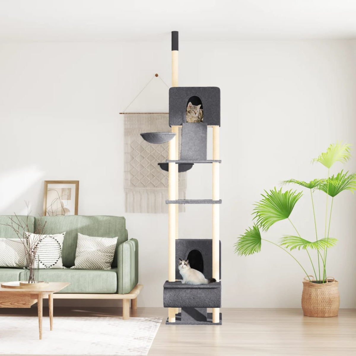 Ein Kratzbaum mit zwei Katzen, eine in einem Versteck, die andere oben. Eine grüne Couch, eine Pflanze und ein gewebter Wandbehang sind ebenfalls sichtbar.