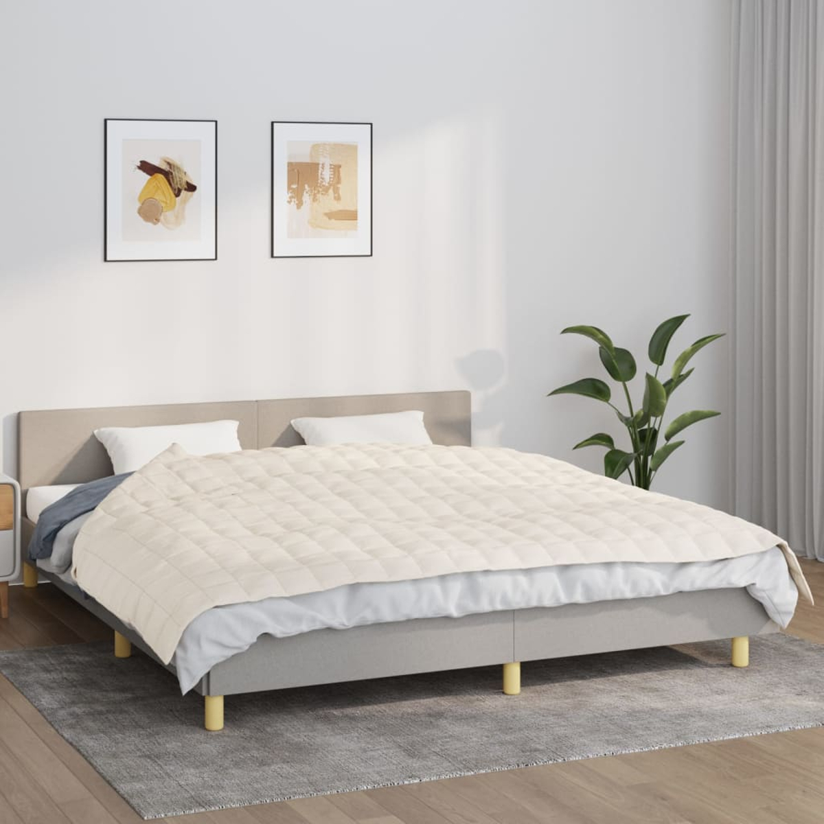 Ein Bett mit beigem Steppbett, Kissen und grauem Kopfteil. Zwei gerahmte Bilder hängen darüber. Eine Pflanze steht daneben.