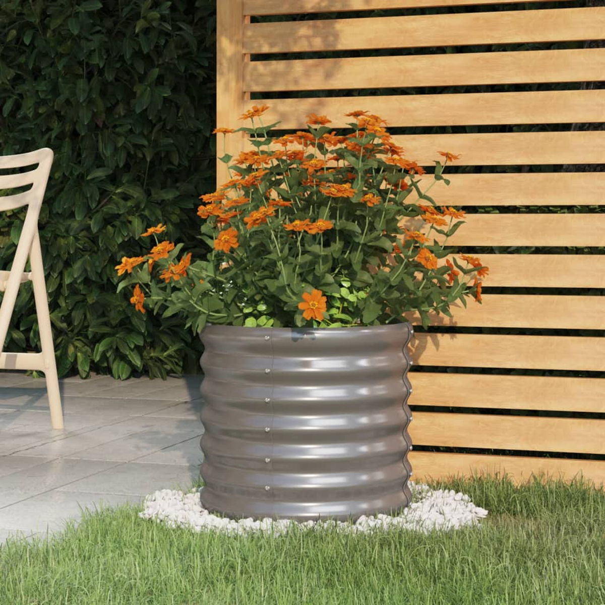 Ein grauer Metallpflanzer mit orangefarbenen Blumen steht auf weißen Steinen. Ein Holzzaun und grünes Gras vervollständigen die Szene.